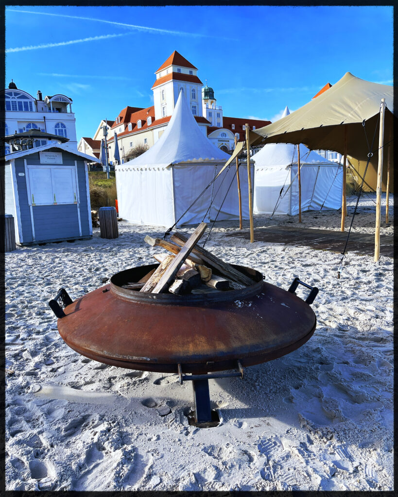 a metallic fire cauldron on the beach. Already filled with some large pieces of wood. Parts of the Binz Kurhotel in the background