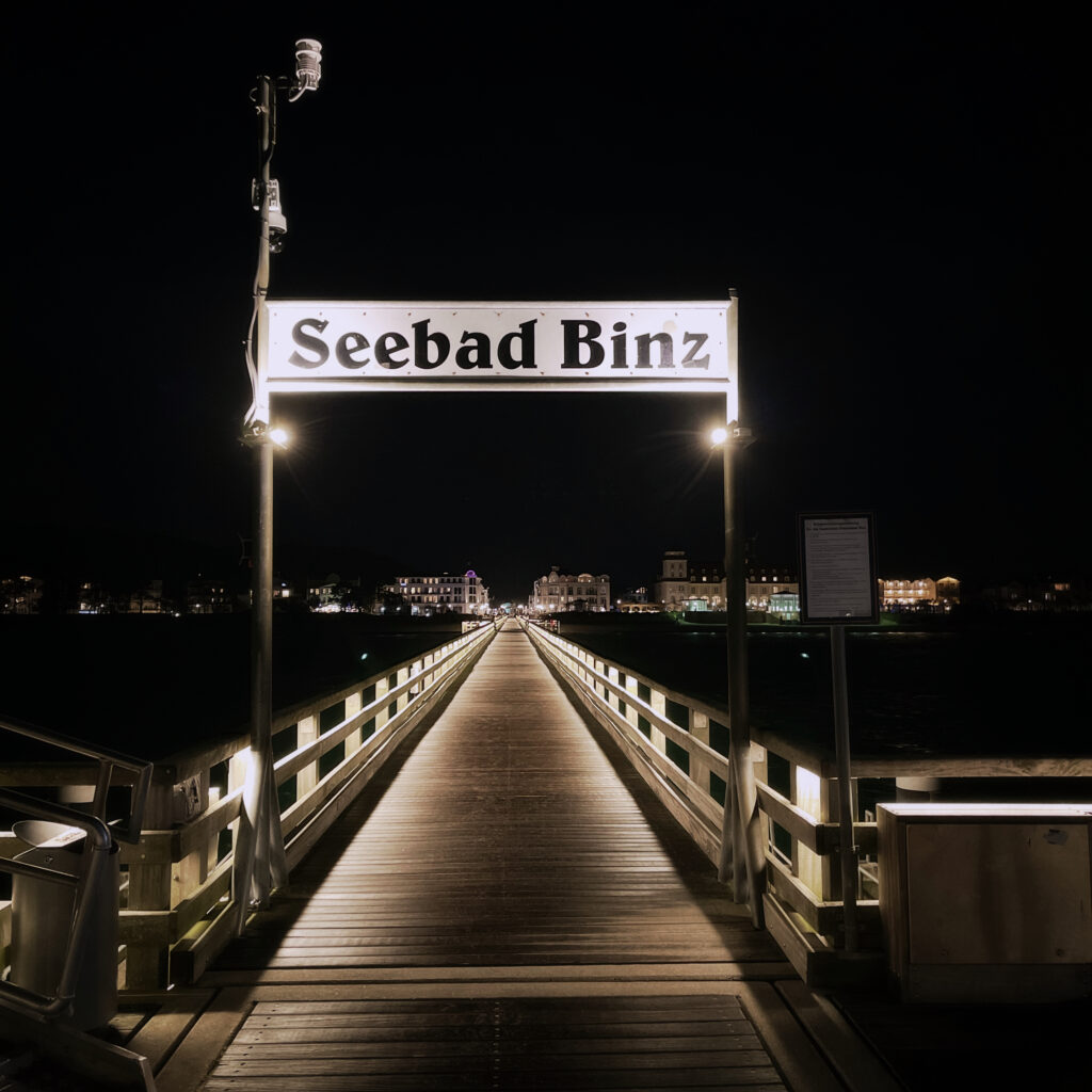 signpost on the sea bridge in Binz. Reading "Seebad Binz". The bridge leads back towards the city and some illuminated buildings can be seen.