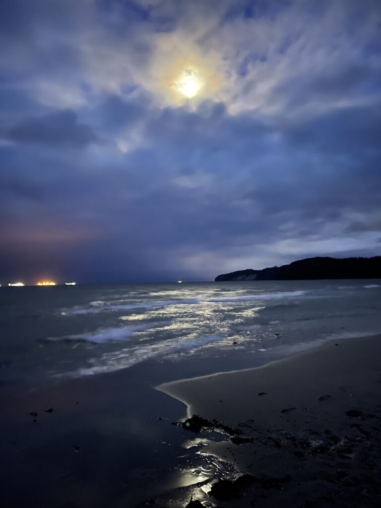 the moon over the baltic sea behind some faint clouds. It reflects in the water and puddles on the shore.