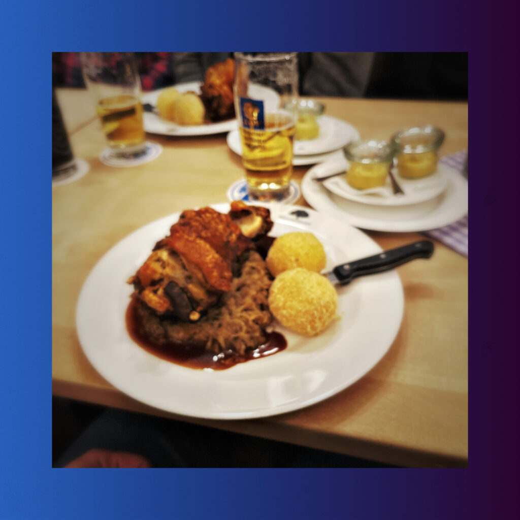 A plate with a crispy pork knuckle, Bavarian sour kraut and dumplings. In the background some beers. 