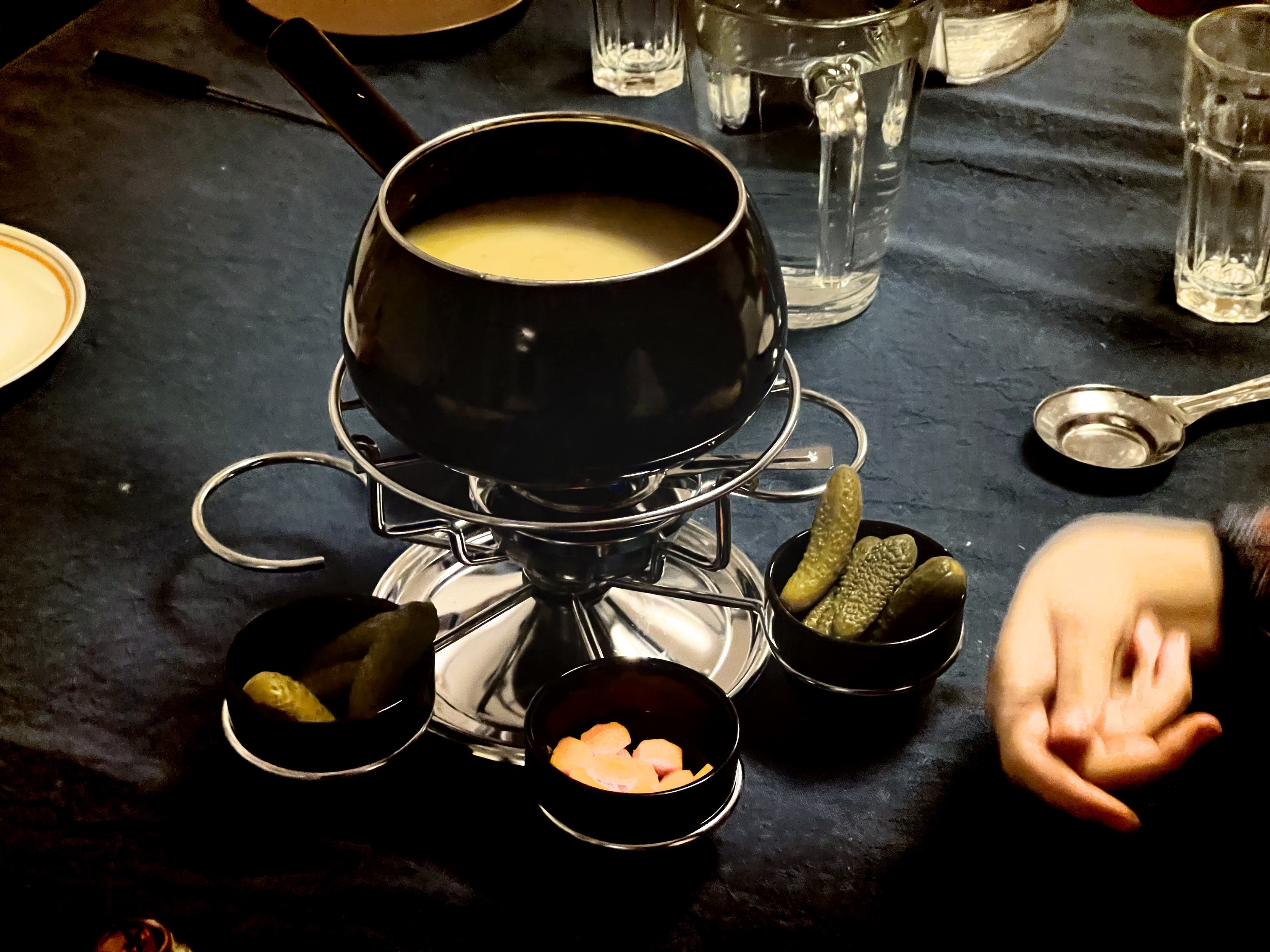 Fondue set on a dining table with a pot of melted cheese, dipping bowls with pickles and other items, and empty glasses. A person's hand is visible to the right.