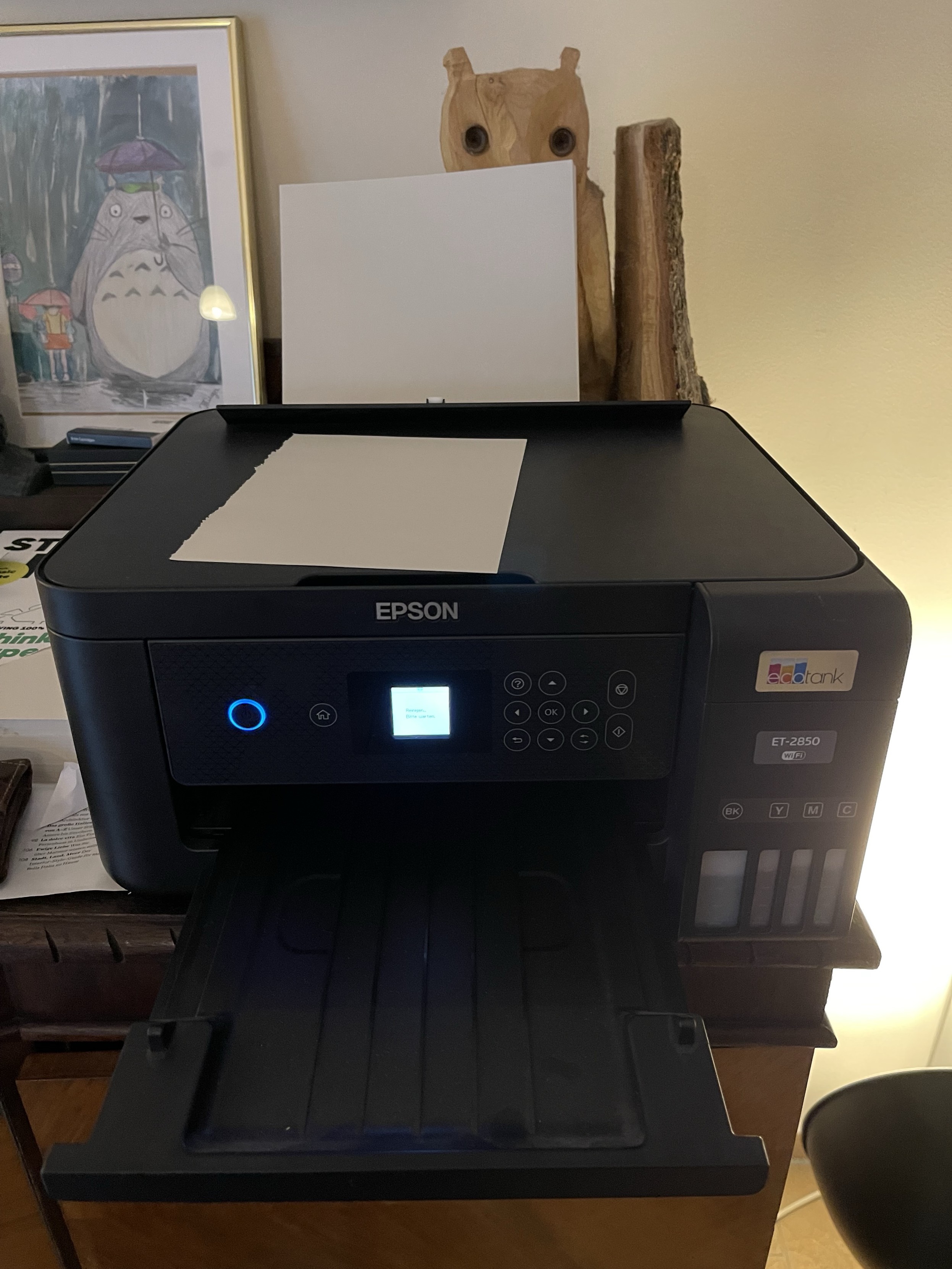 An Epson ink tank printer on a desk with a sheet of paper on the scanner bed, next to decorative items including a framed picture of a character and a wooden figure.