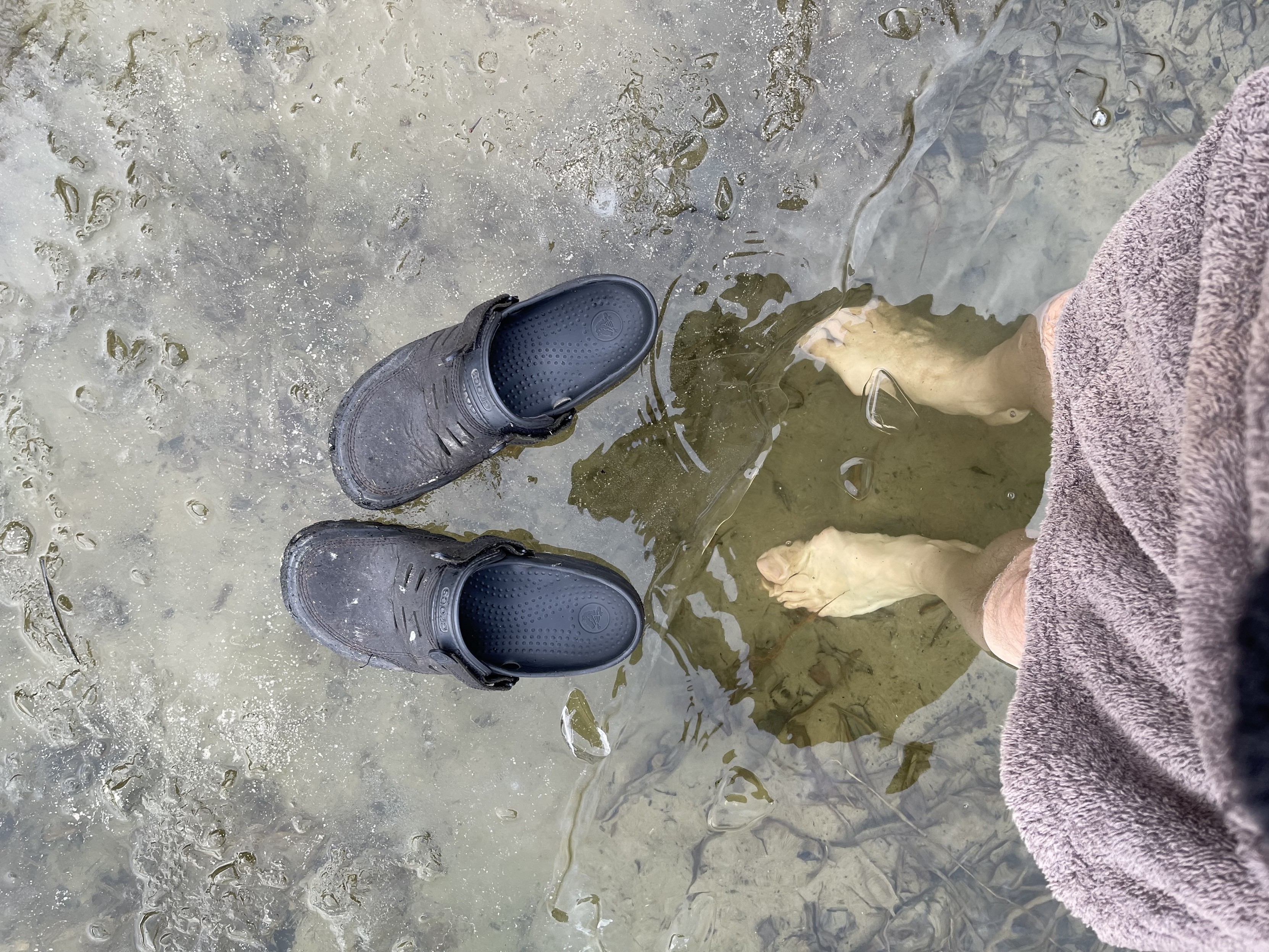 Person standing in a hole in the ice of a lake with bare feet next to a pair of grey shoes on the ice. 