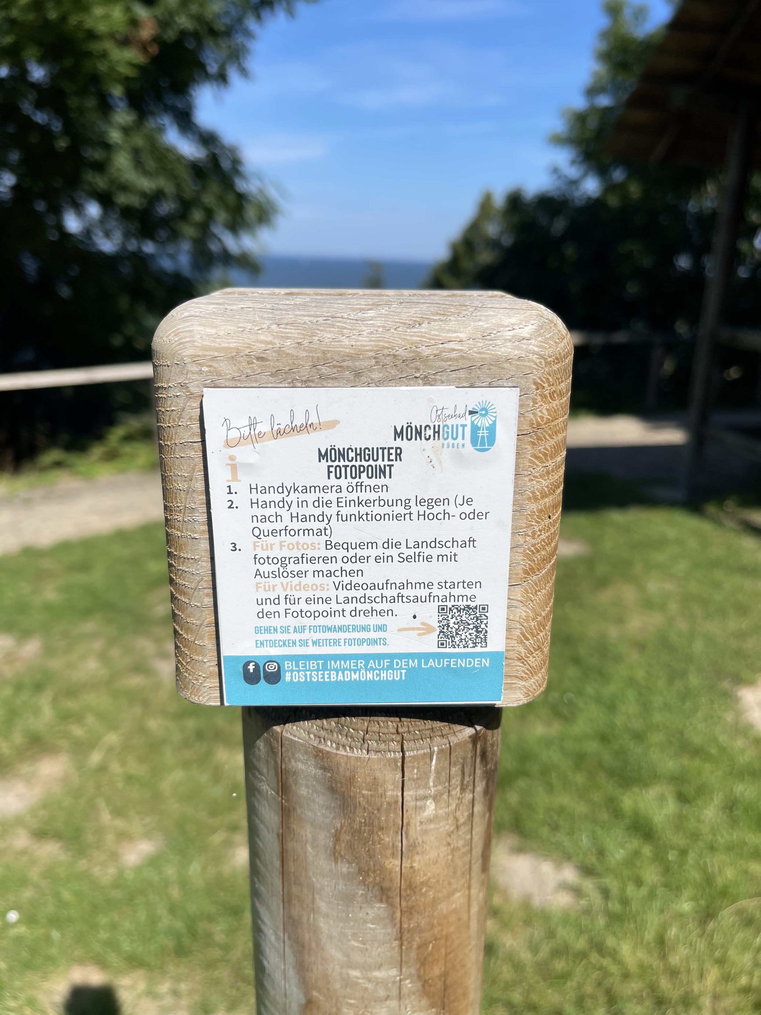 Schild mit Anweisungen für einen Fotopunkt in Ostseebad Mönchgut, Deutschland. Das Schild erklärt, wie man die Telefonkamera öffnet, das Telefon positioniert und Fotos oder Videos aufnimmt. Bäume und ein weit entferntes Gewässer sind sichtbar in