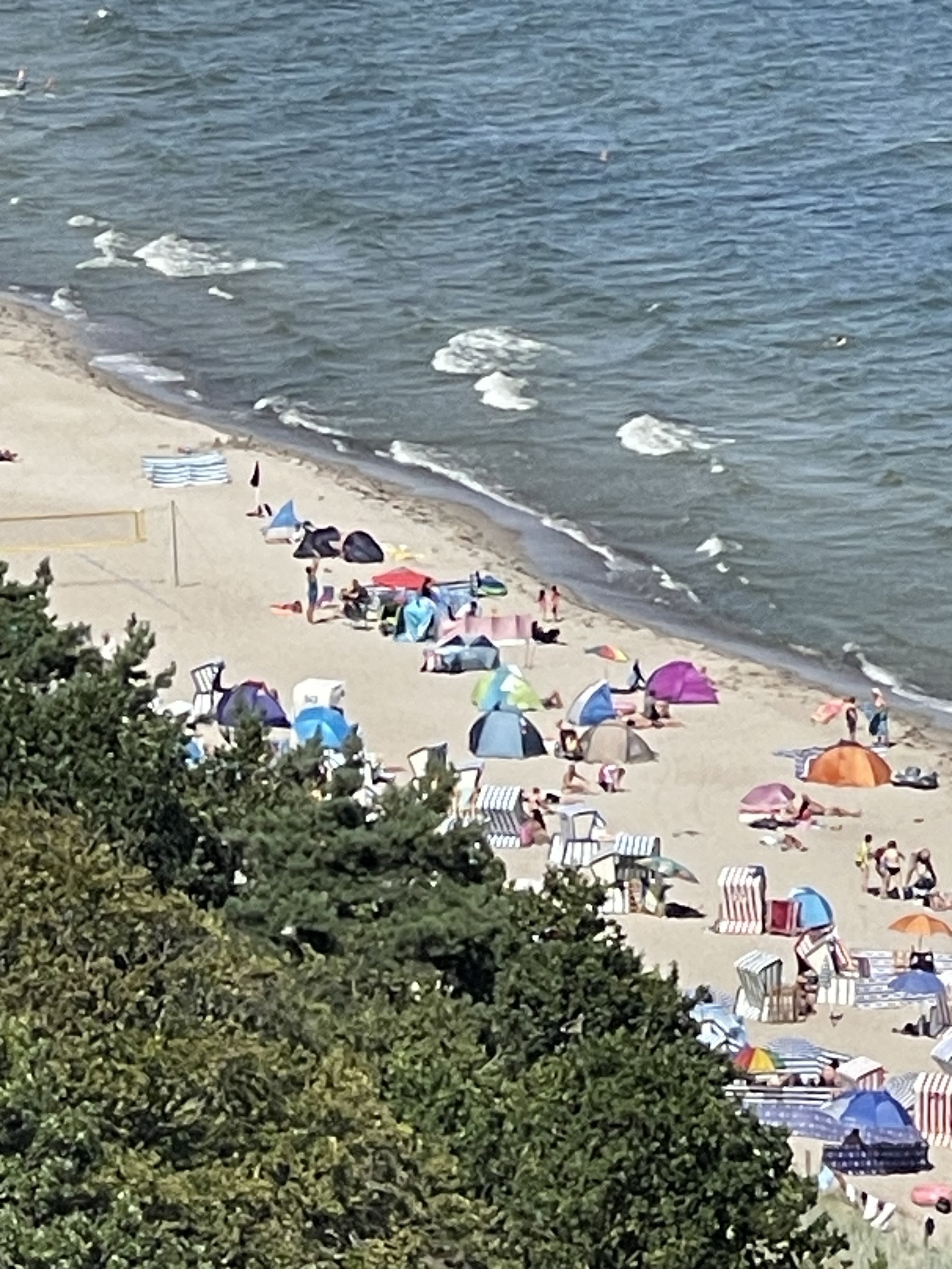 Eine Strandszene mit Menschen, die sich unter bunten Sonnenschirmen und Zelten entspannen, zusammen mit einigen in der Nähe des Wassers. Wellen und Bäume sind ebenfalls sichtbar.