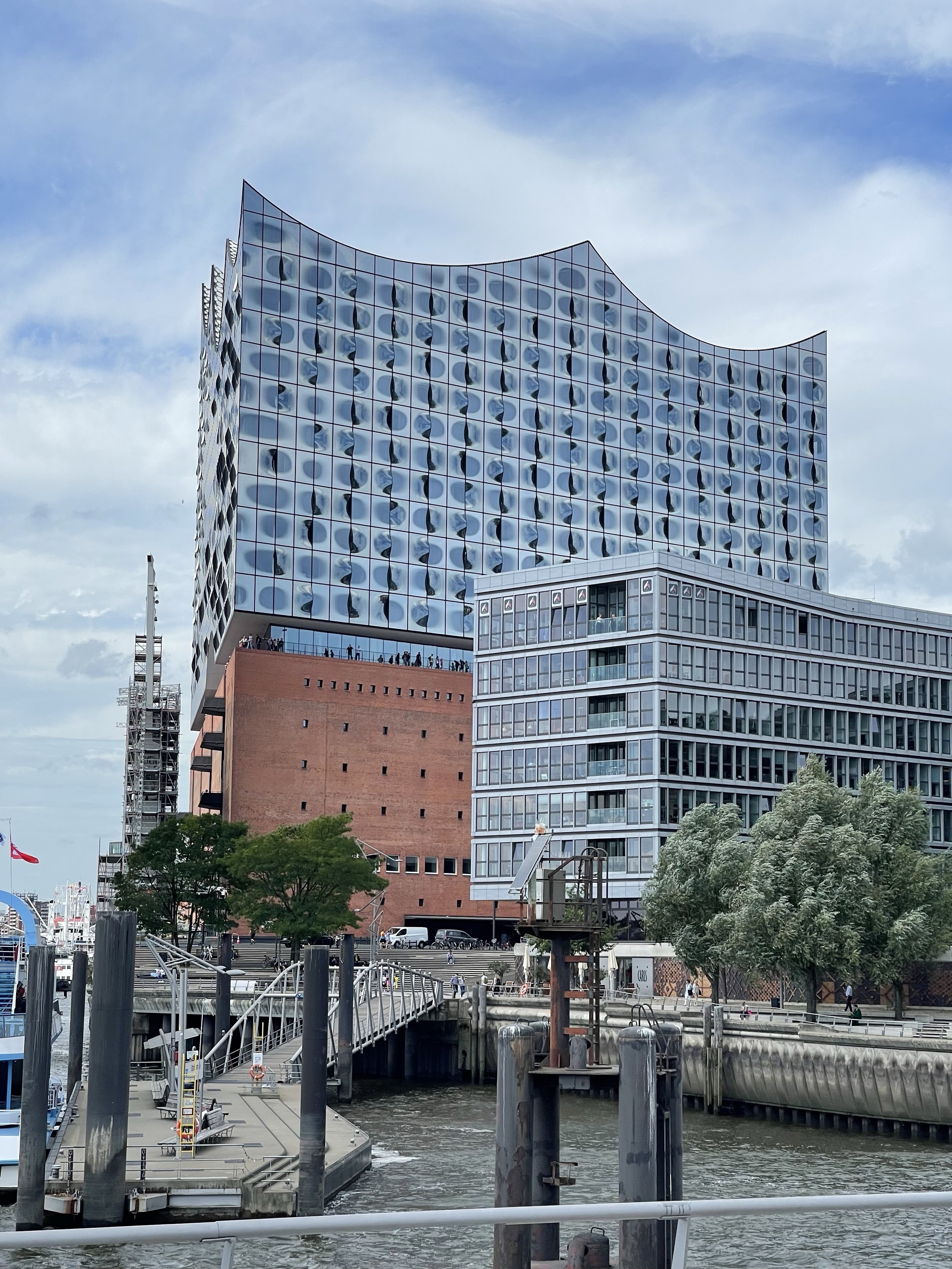 A modern architectural building with a curved glass facade is prominently featured, alongside a brick structure. The scene includes a riverside promenade with people, trees, and boats in the water, under a cloudy sky.
The building is the Elbphilharmonie in Hamburg