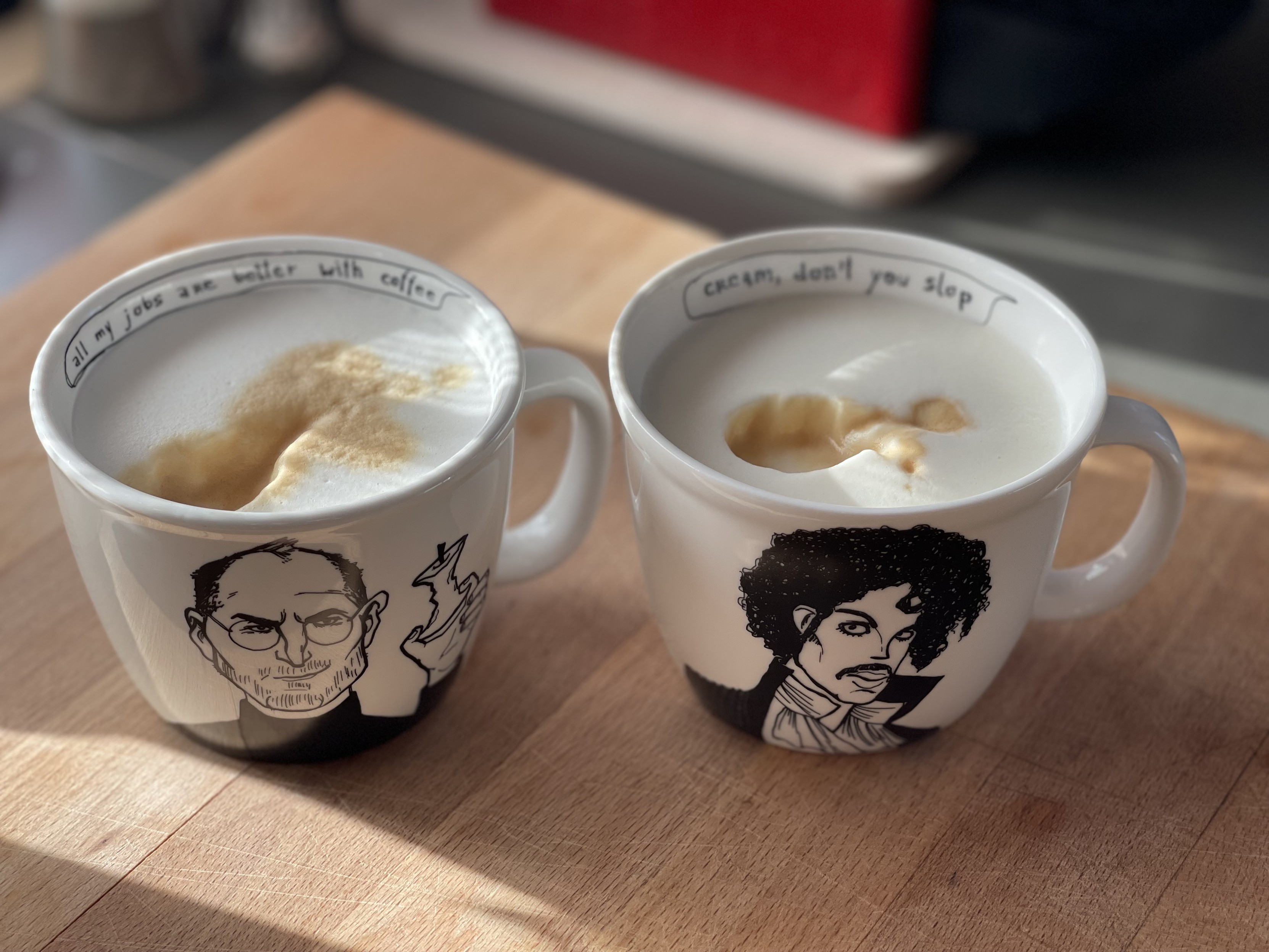 Two coffee mugs with illustrated portraits on them and text, sitting on a wooden surface with sunlight casting shadows.
Left illustration is Steve Jobs holding an Apple corpse. Text inside the mug seam is: all my jobs are better with coffee. 

Right side is Prince and the text on the seam is: Cream, don't you slop 