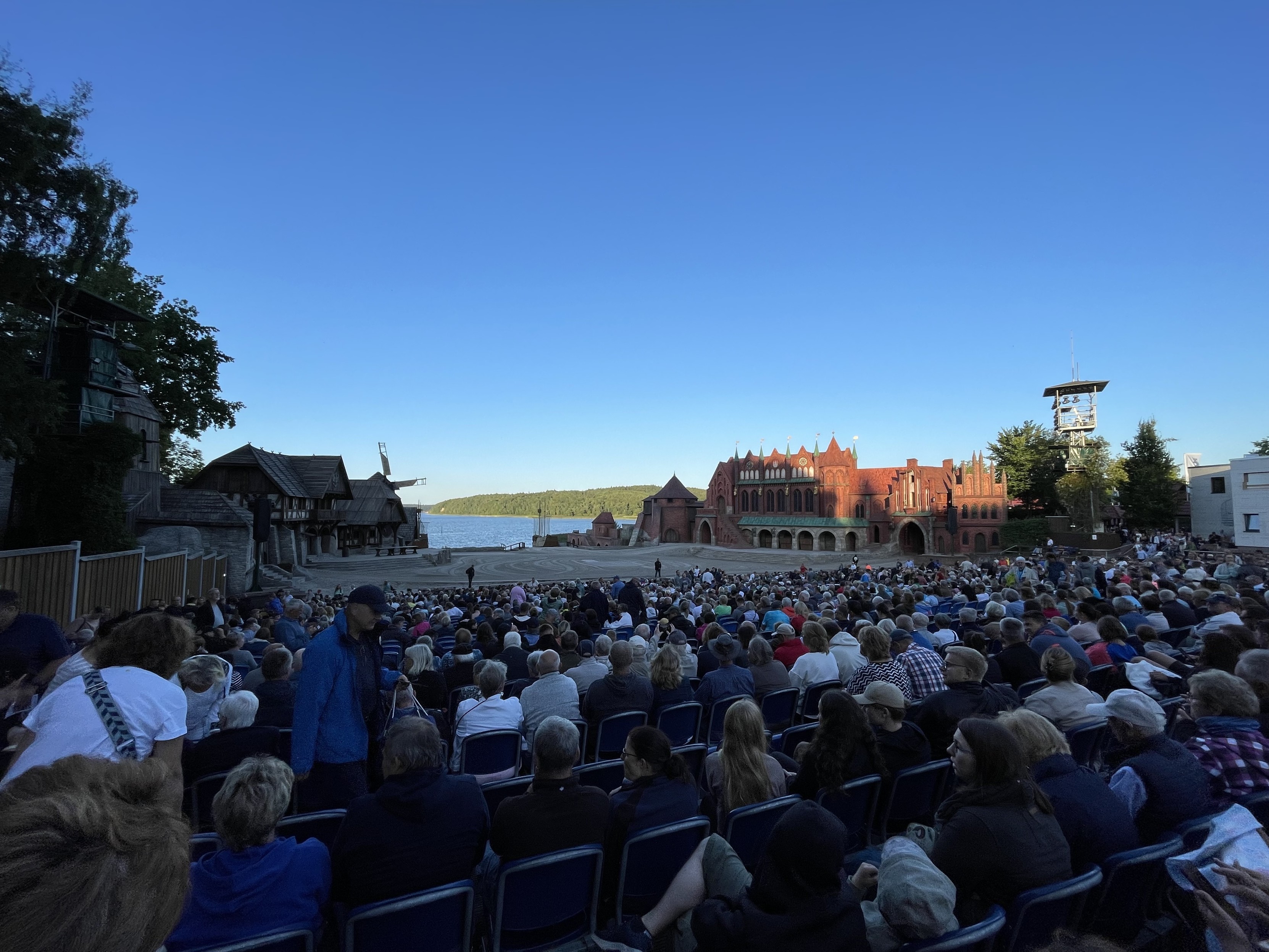 Outdoor-Theater mit einem großen Publikum, das auf eine Bühne blickt, die wie eine mittelalterliche Stadt aussieht, mit Gebäuden und einer Windmühle im Hintergrund unter einem klaren blauen Himmel.