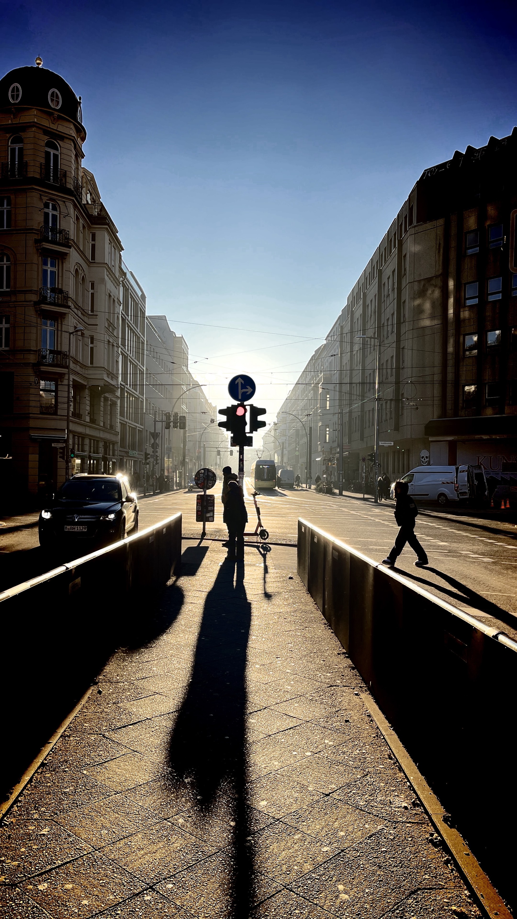 Urban street scene with backlight creating silhouettes of people and long shadows, traffic signal, tram, and architecture, under a clear sky.