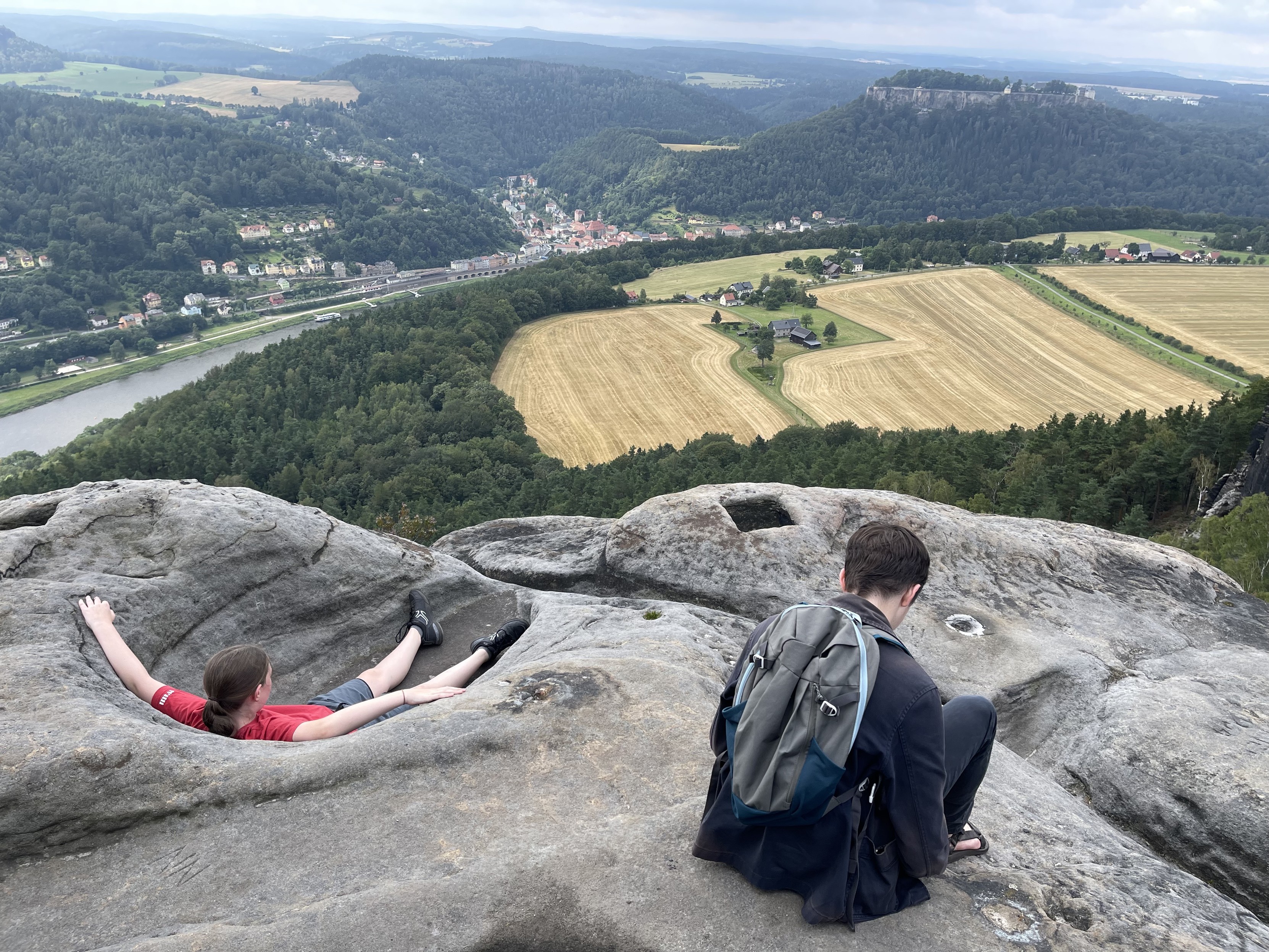 Two people on a rocky cliff overlooking a scenic landscape with fields, forests, a river, and distant hills. One person is lying down, while the other sits with a backpack.