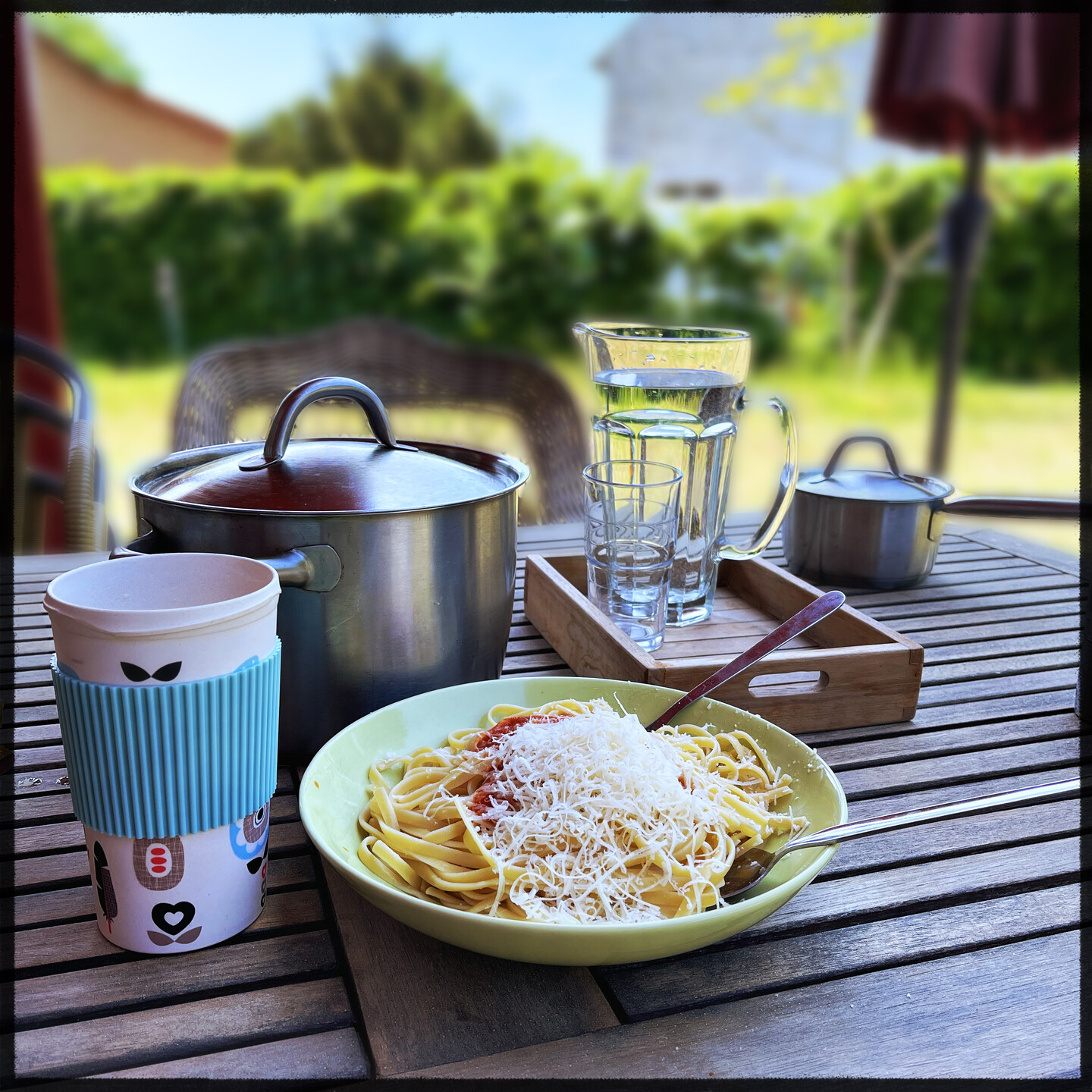 A lunch setup in a wooden table outside. In front a plate with linguine pasta, tomato sauce and Parmesan cheese on top. Behind in the left is a large cooking pot with lid. Behind middle is a small wooden Tablet with a glass and a carafe of water. In the far background a green hedge some house and blue sky. All blurry