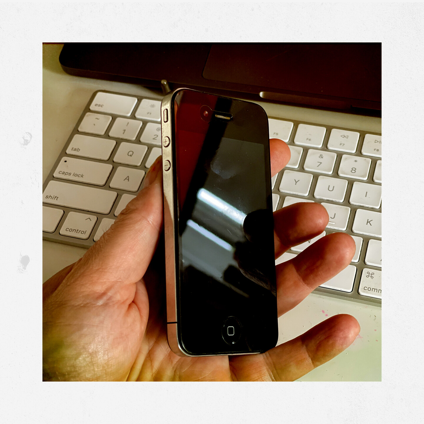 An Apple iPhone 4 held by a left hand above an Apple keyboard. 