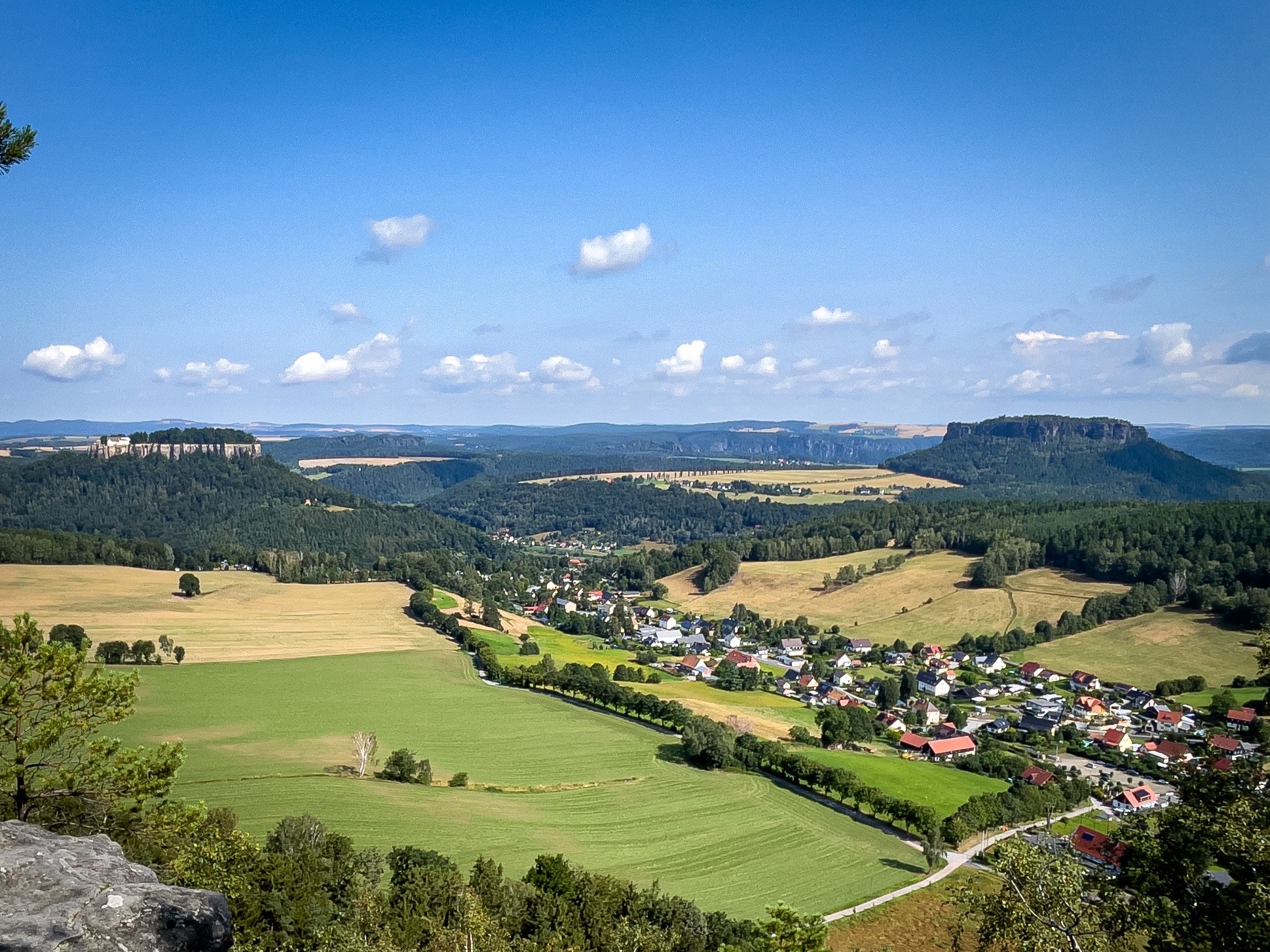 Malerische Landschaft mit sanften Hügeln, einem kleinen Dorf, offenen Feldern und dichten Wäldern, unter einem klaren blauen Himmel mit verstreuten Wolken.
Links hinten die Festung Königstein, rechts hinten der Lilienstein 