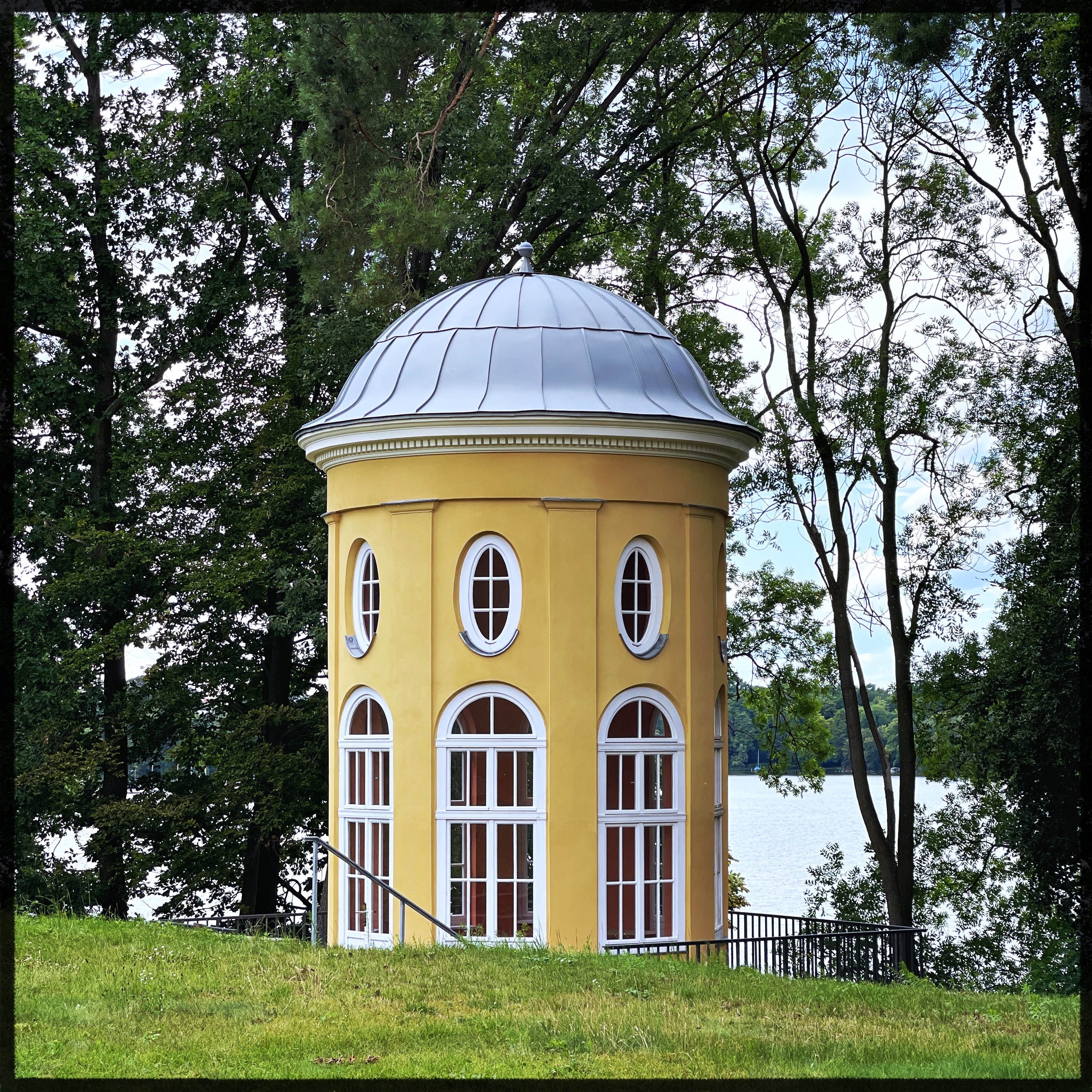 Eine kleine gelbe Rotunde mit einem silbernen Kupeldach, zwischen Bäumen und mit Blick auf einen See, mit gewölbten Fenstern sowohl auf dem Boden als auch auf der oberen Ebene.