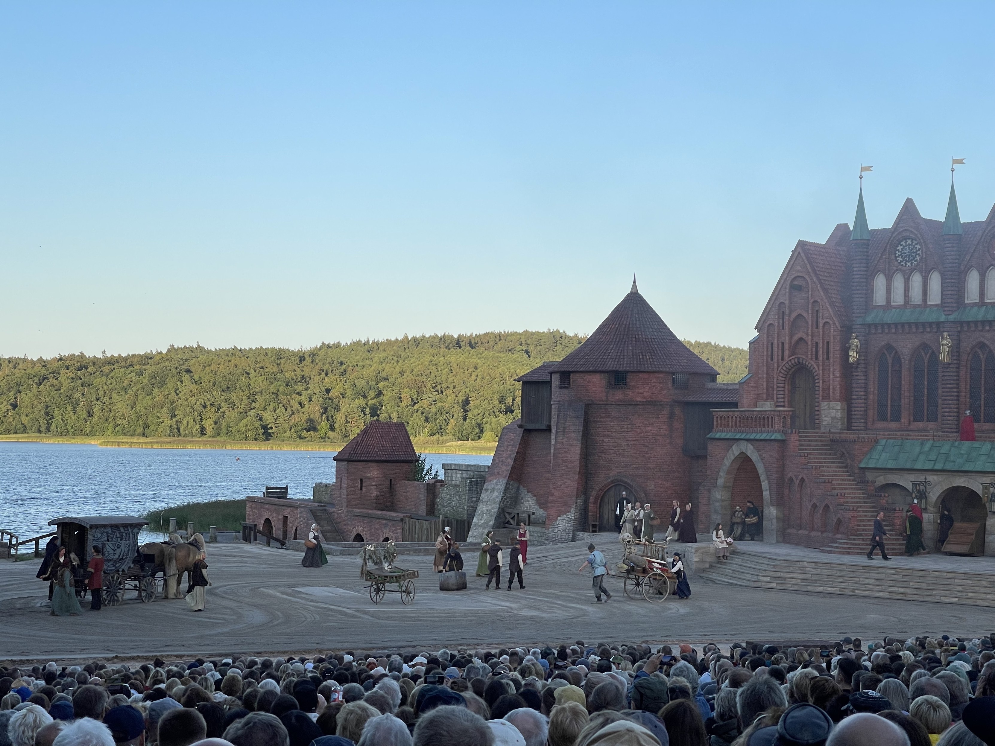 Theateraufführung im Freien mit einem großen Publikum, einer Bühne, die einem alten Schloss ähnelt, und Schauspielern in zeitlichen Kostümen in der Nähe eines malerischen Sees.