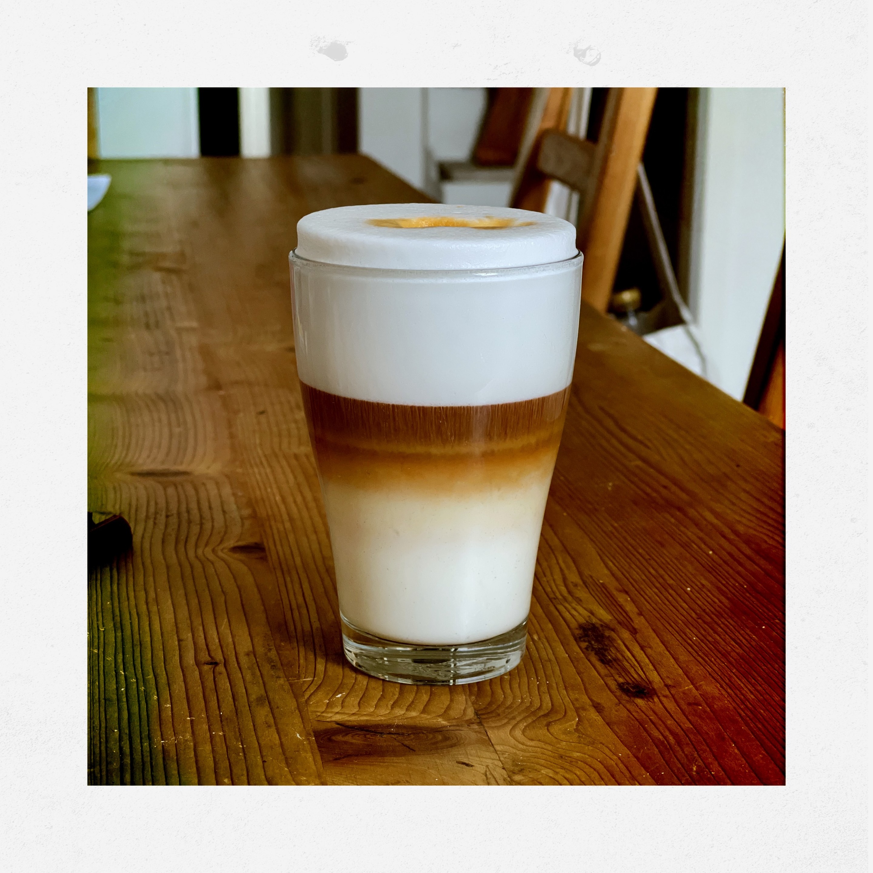 Polaroid style pictures of a glass of foamed milk with a brown/red liquid floating in the middle of the glass. The glass stands on a wooden table. 