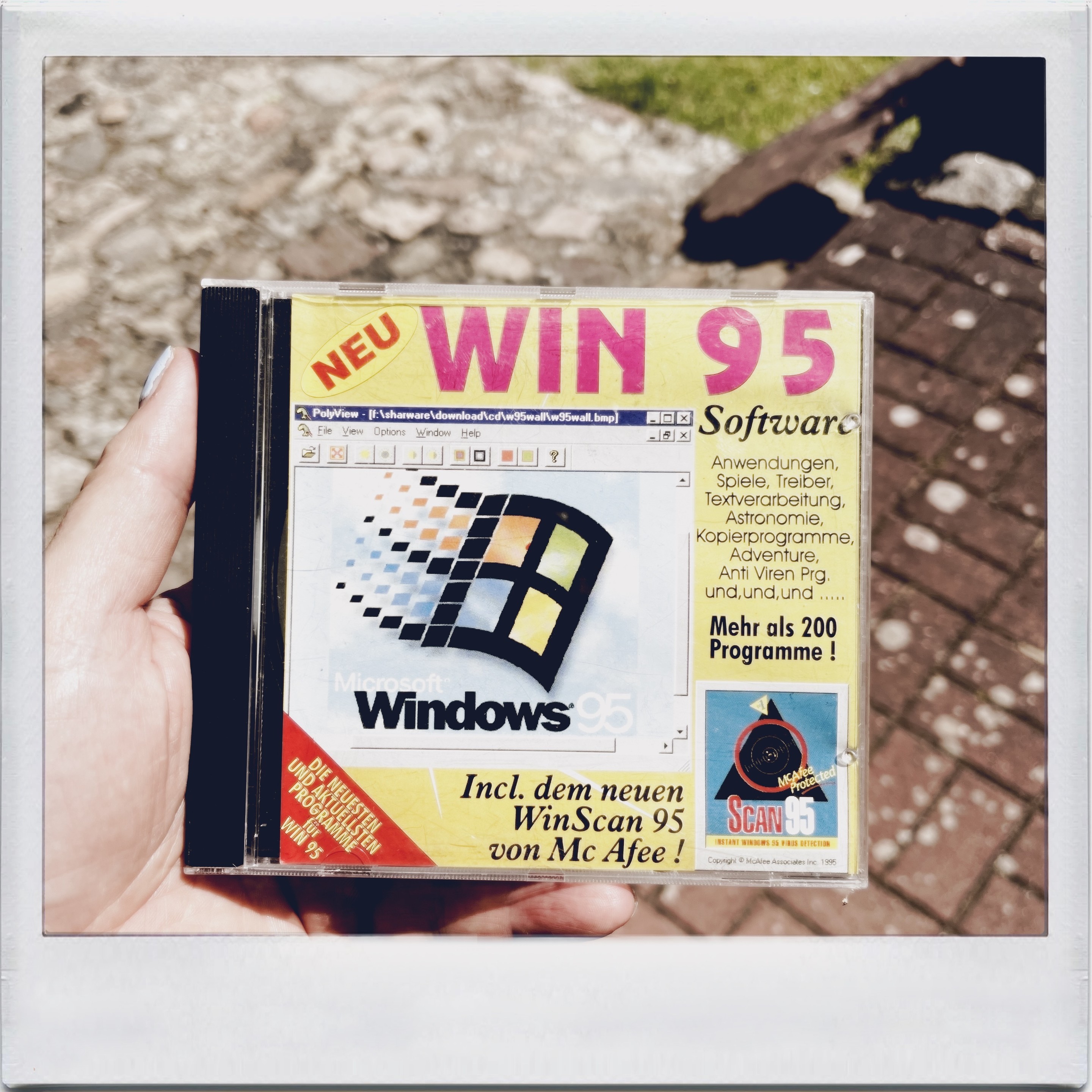 A hand holding a Windows 95 software CD package with German text, showing the Windows logo and advertising more than 200 programs, including WinScan 95 by McAfee. The background includes a stone path and grass.