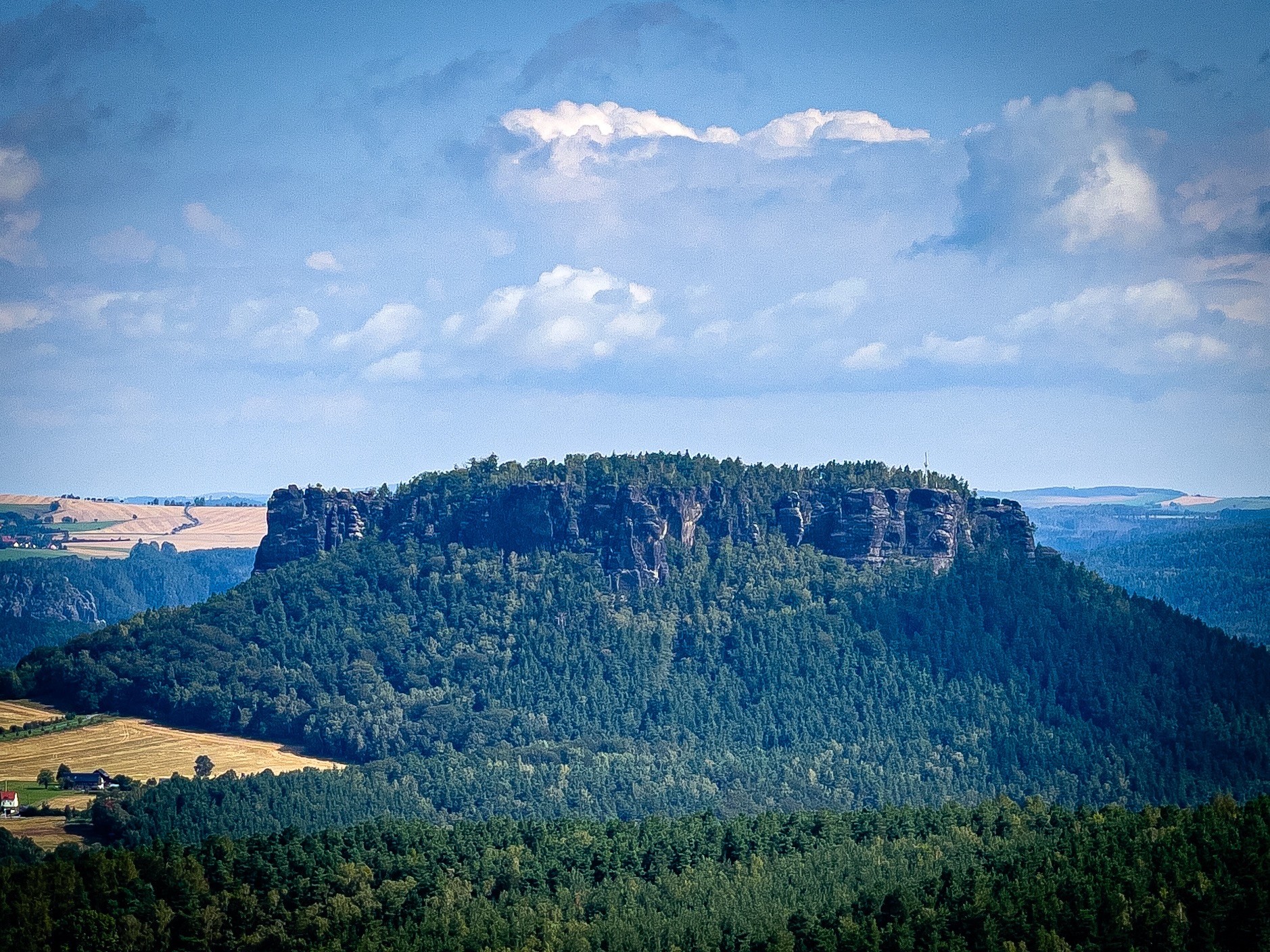 Eine flache Mesa, die mit dichtem grünem Wald bedeckt ist, vor dem Hintergrund von sanften Hügeln und einem Himmel, der mit verstreuten Wolken gefüllt ist.
Es ist der Lilienstein in der sächsischen Schweiz 