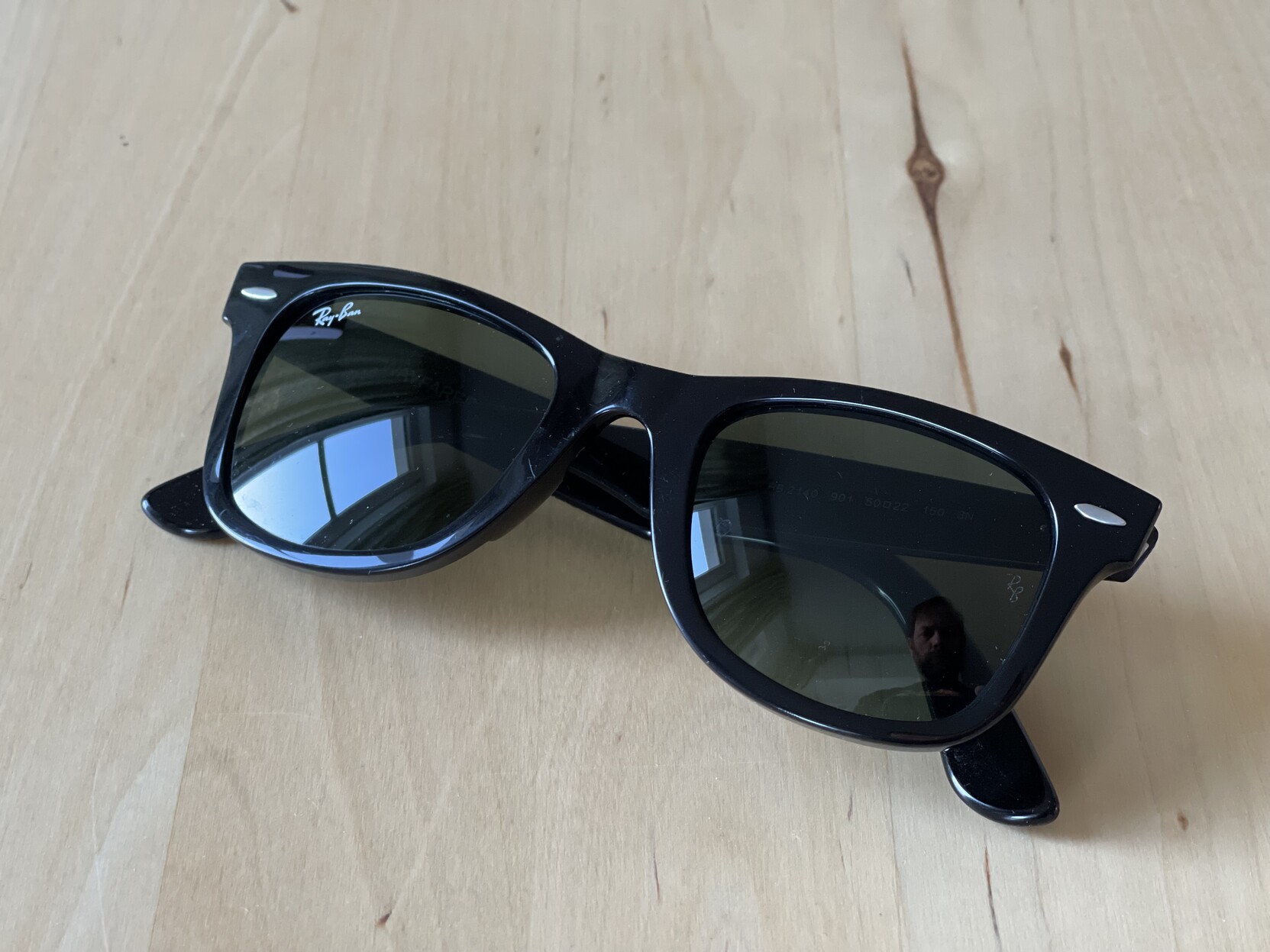 Eine Sonnenbrille: Ray Ban New Wayfarer in schwarz. Liegt auf einem Tisch aus hellem Holz. 