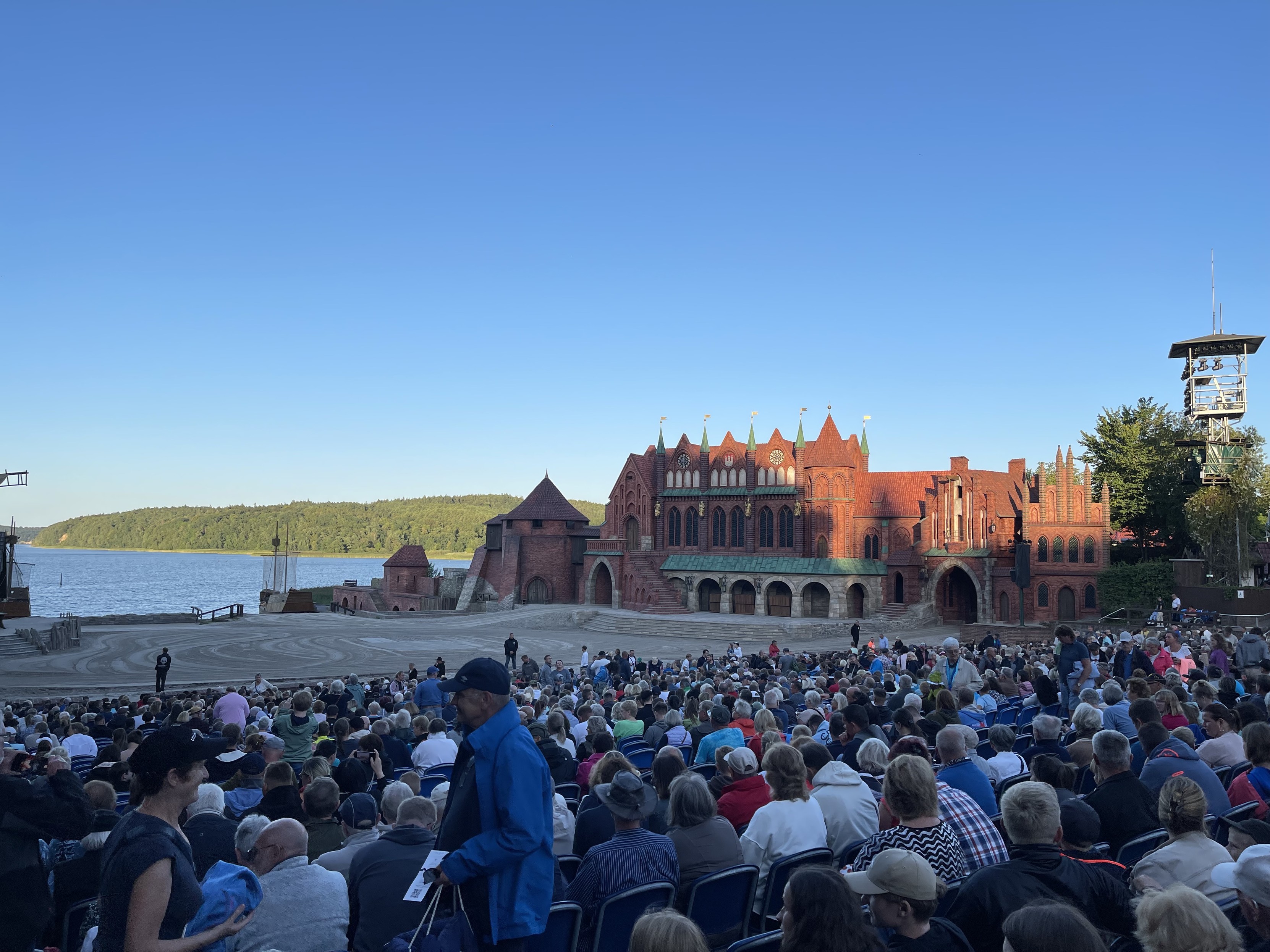 Open-Air-Theater mit einem großen Publikum, das vor einer Bühne steht, die einem historischen roten Backsteingebäude ähnelt und an einem See liegt, der von grünen Hügeln umgeben ist.