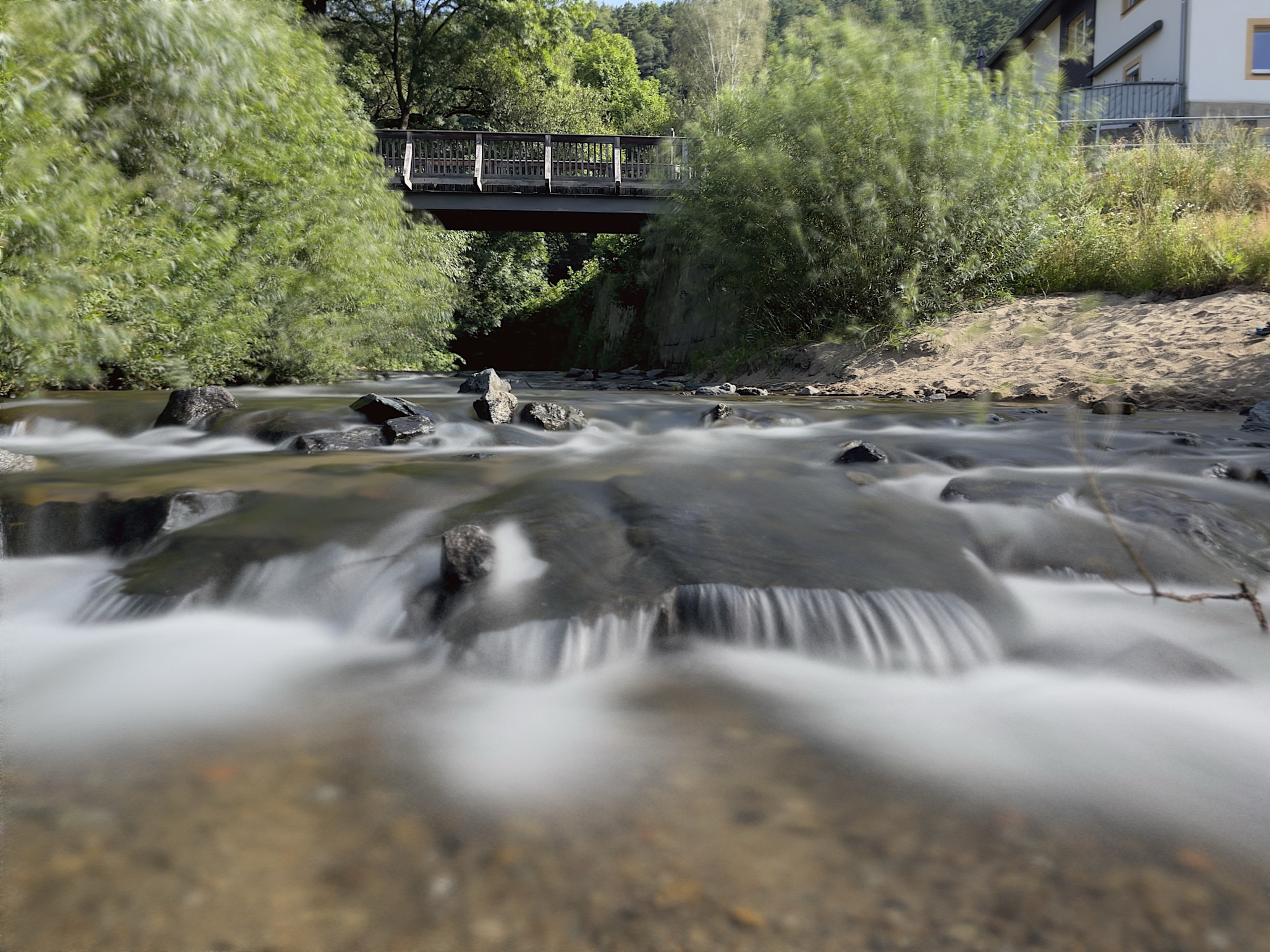 A serene creek flows over rocks under a pedestrian bridge, surrounded by lush greenery and a building visible on the right side.