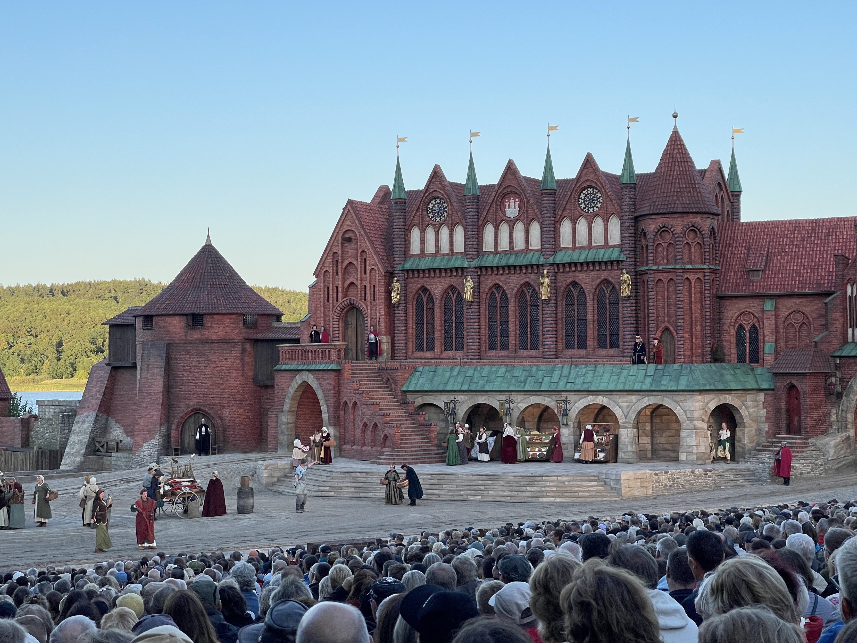 Theateraufführung im Freien mit Schauspielern in mittelalterlichen Kostümen auf einer Bühne, die einem Schloss ähnelt, mit einem großen Publikum, das zuschaut.