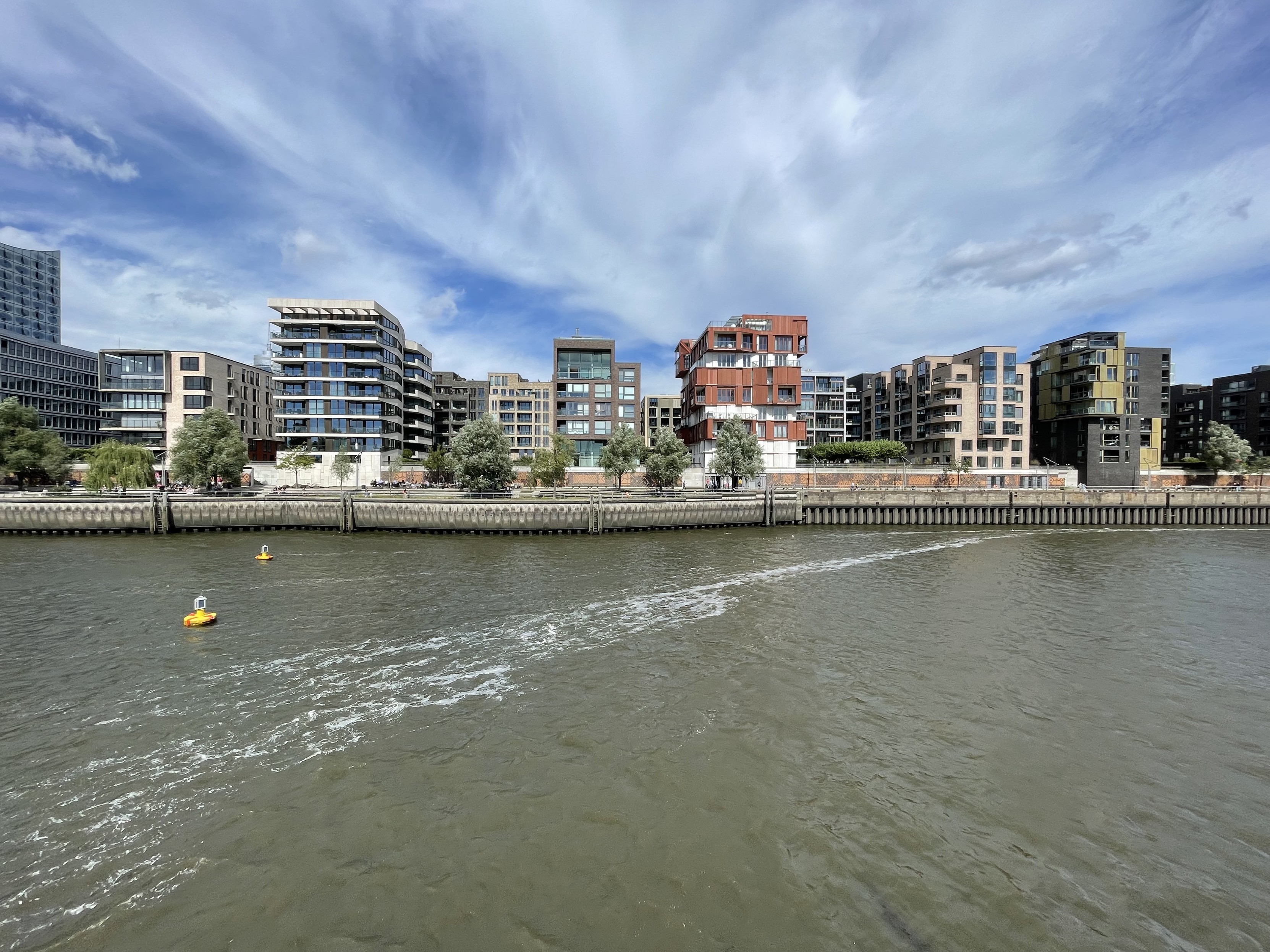 A view of modern buildings along a riverbank, featuring various architectural styles and greenery. Two yellow buoys float in the water, with gentle waves and a partly cloudy sky in the background.