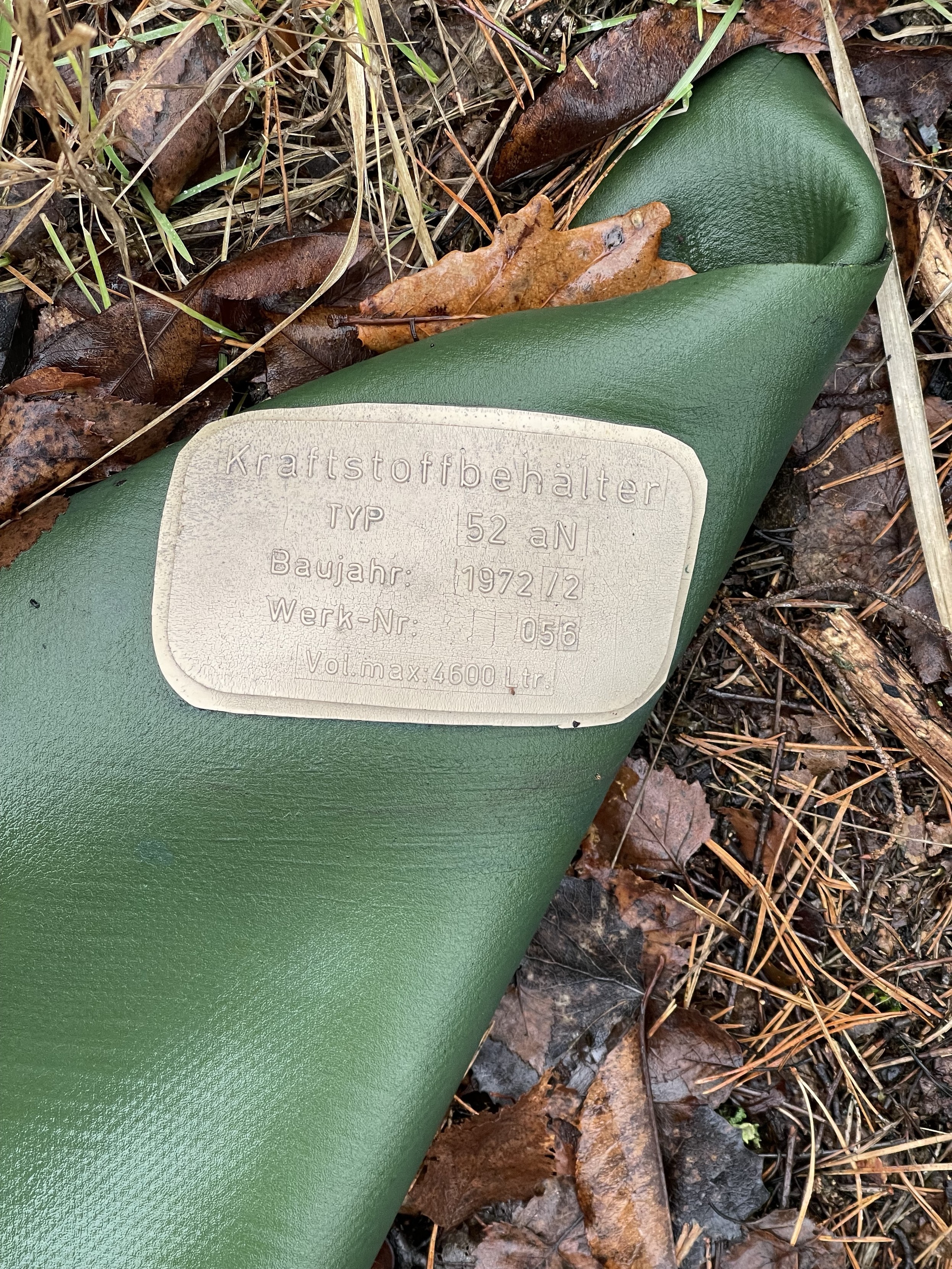 Green cylindrical object with a label in German lying on forest ground with dry leaves. This is a remainder of Soviet army. The label reads that it was a container for fuel. Build in 1972. 
