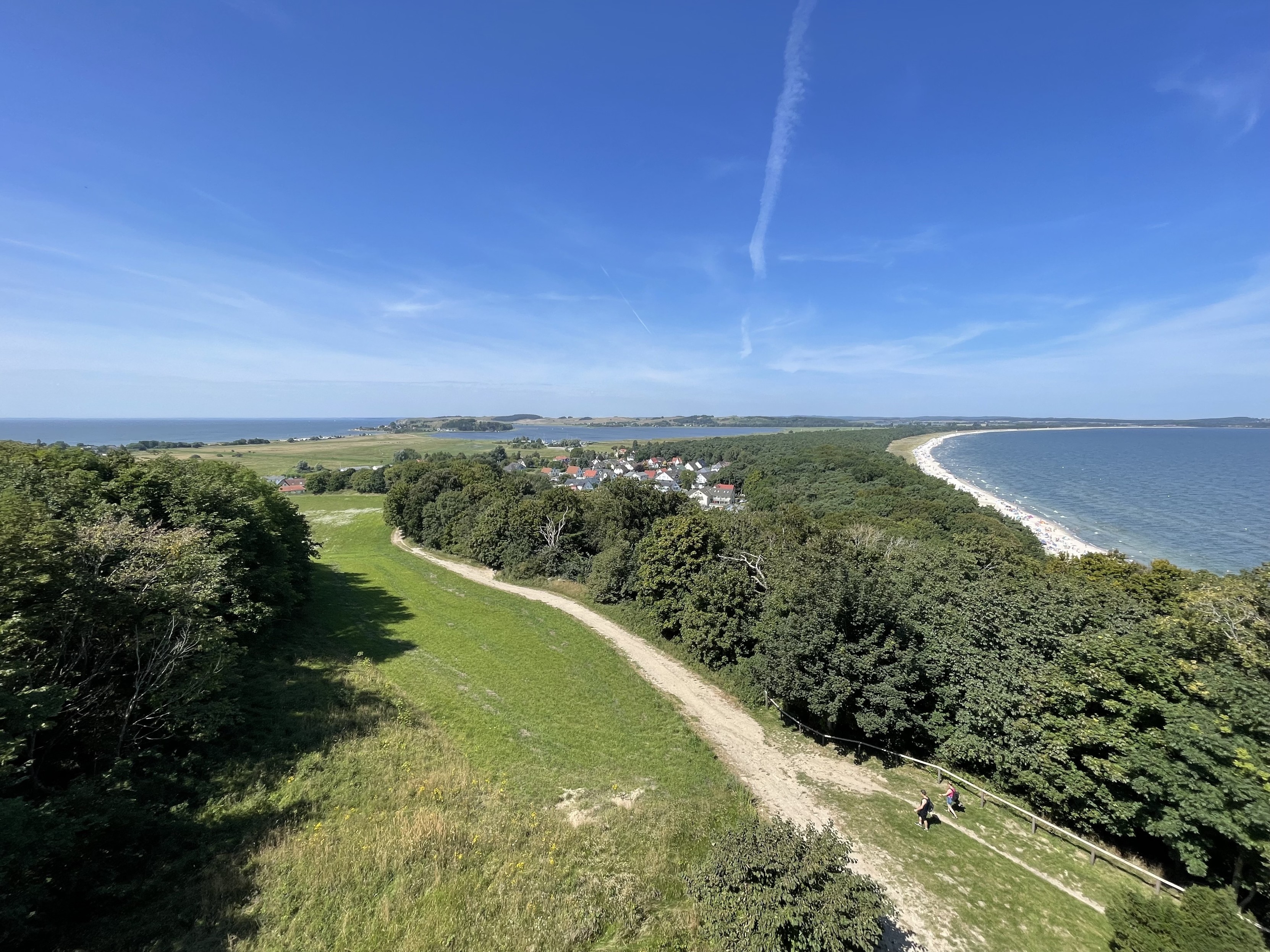 Luftaufnahme einer malerischen Landschaft mit einem Küstendorf, umgeben von Grün, mit einem Strand entlang der Küste und dem Ozean im Hintergrund unter einem klaren blauen Himmel.