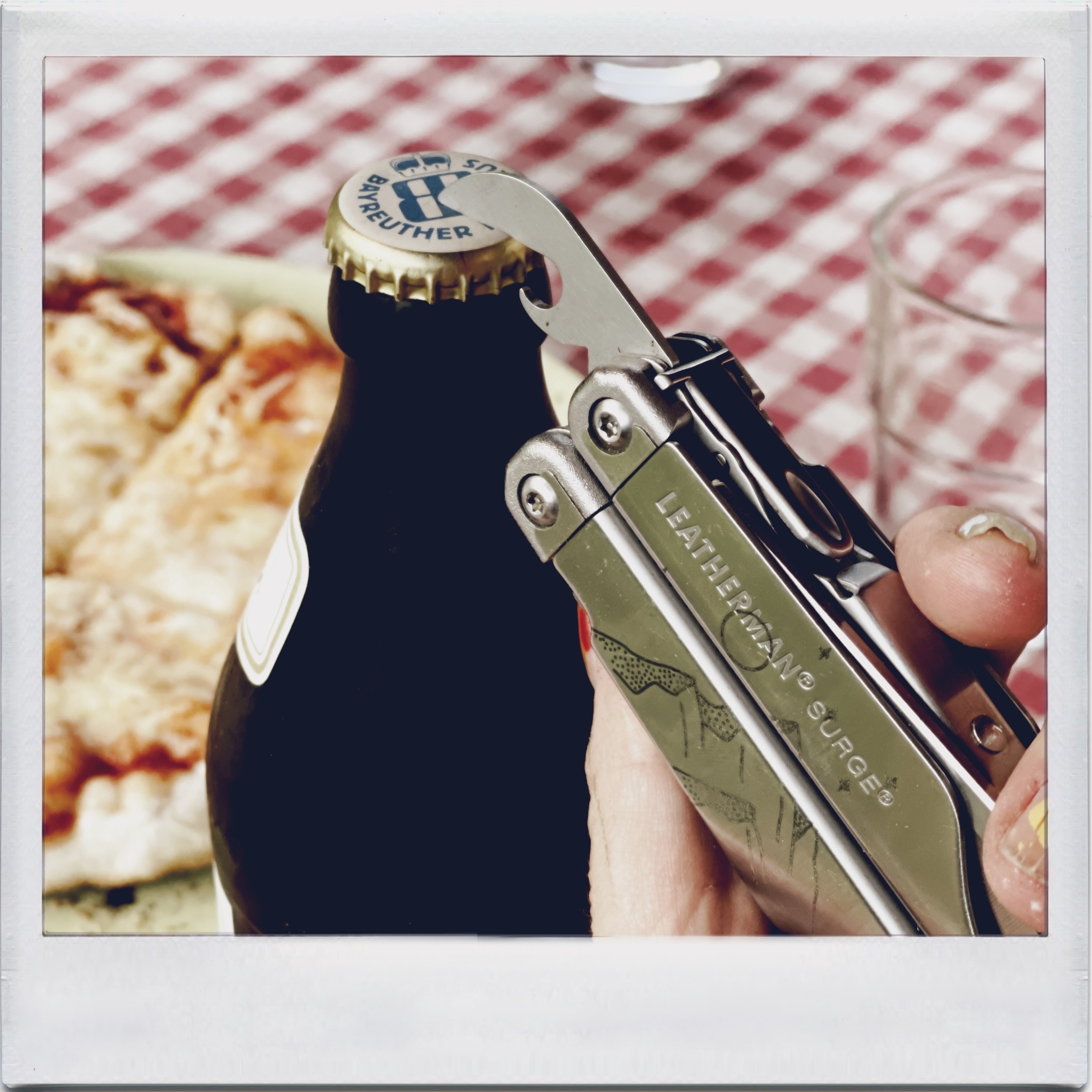 A hand holding a multi-tool is using its bottle opener to pop the cap off a dark glass bottle, with a pizza in the background and a checkered tablecloth.