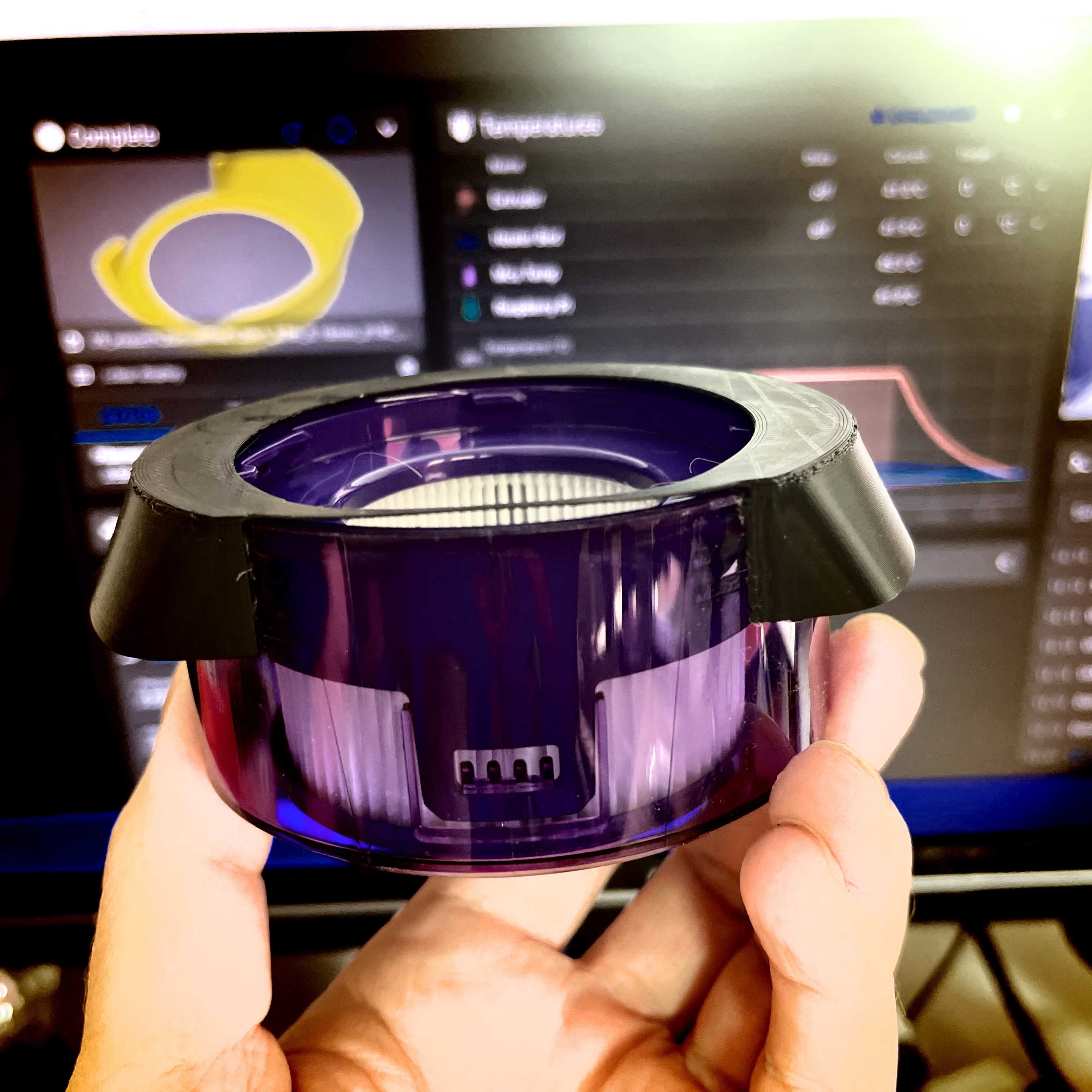 A person holding a Dyson motor filter for vacuum cleaner with a 3D printed lid/air diversion with a blurred computer screen displaying a graphical interface in the background.