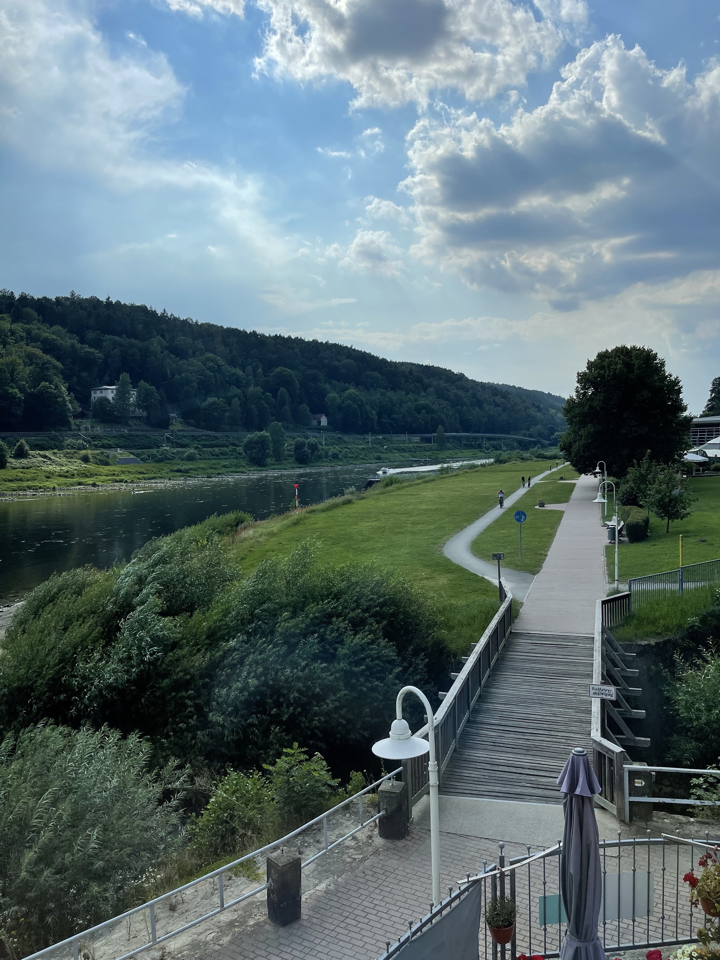 A scenic riverside pathway with greenery, a boardwalk, and a lamppost. The sky is partly cloudy with sunlight breaking through. The path follows the curve of the river, with trees and hills in the background.