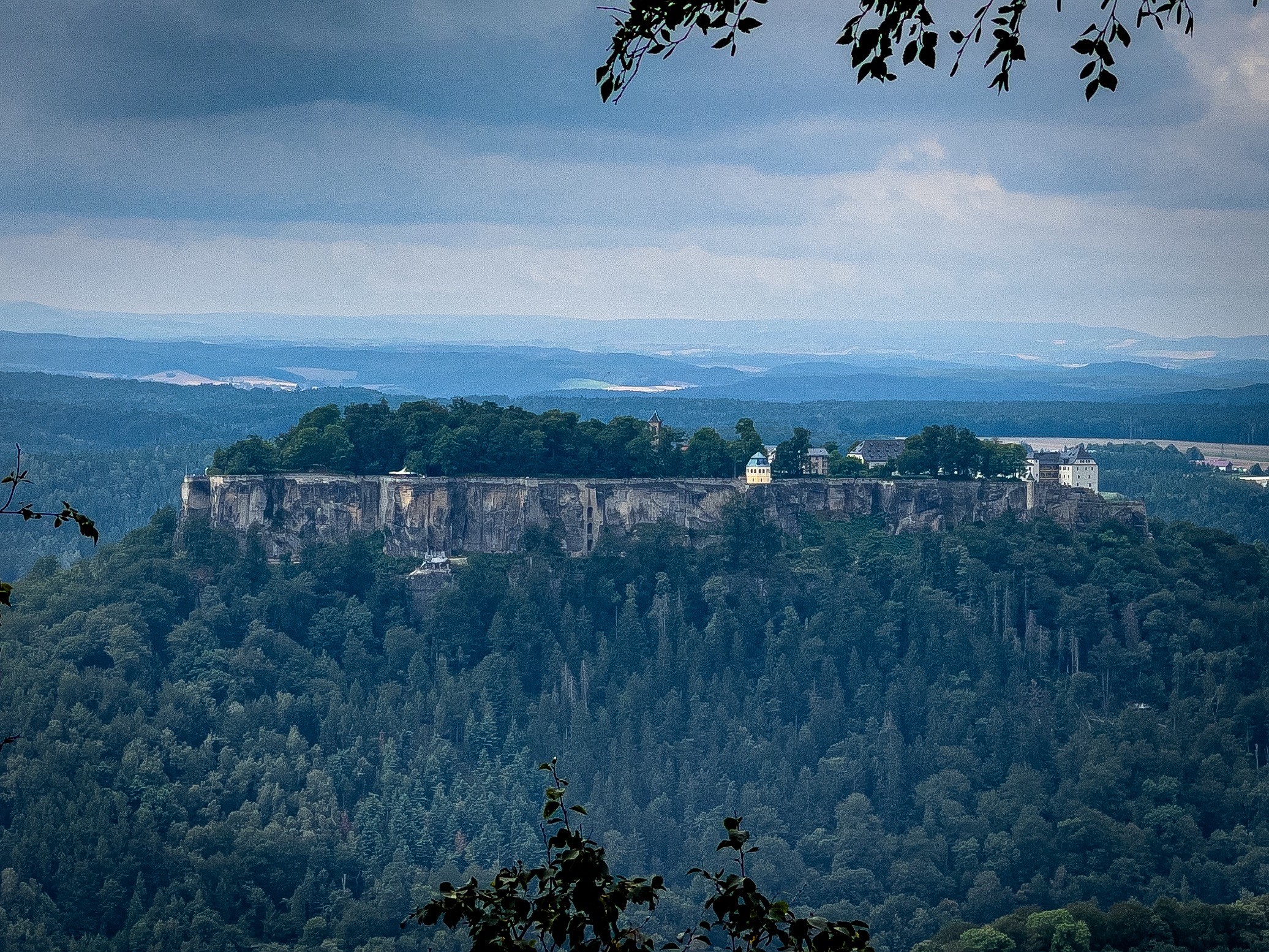 A distant view of a fortress built on a forested plateau, surrounded by lush greenery and under a cloudy sky.