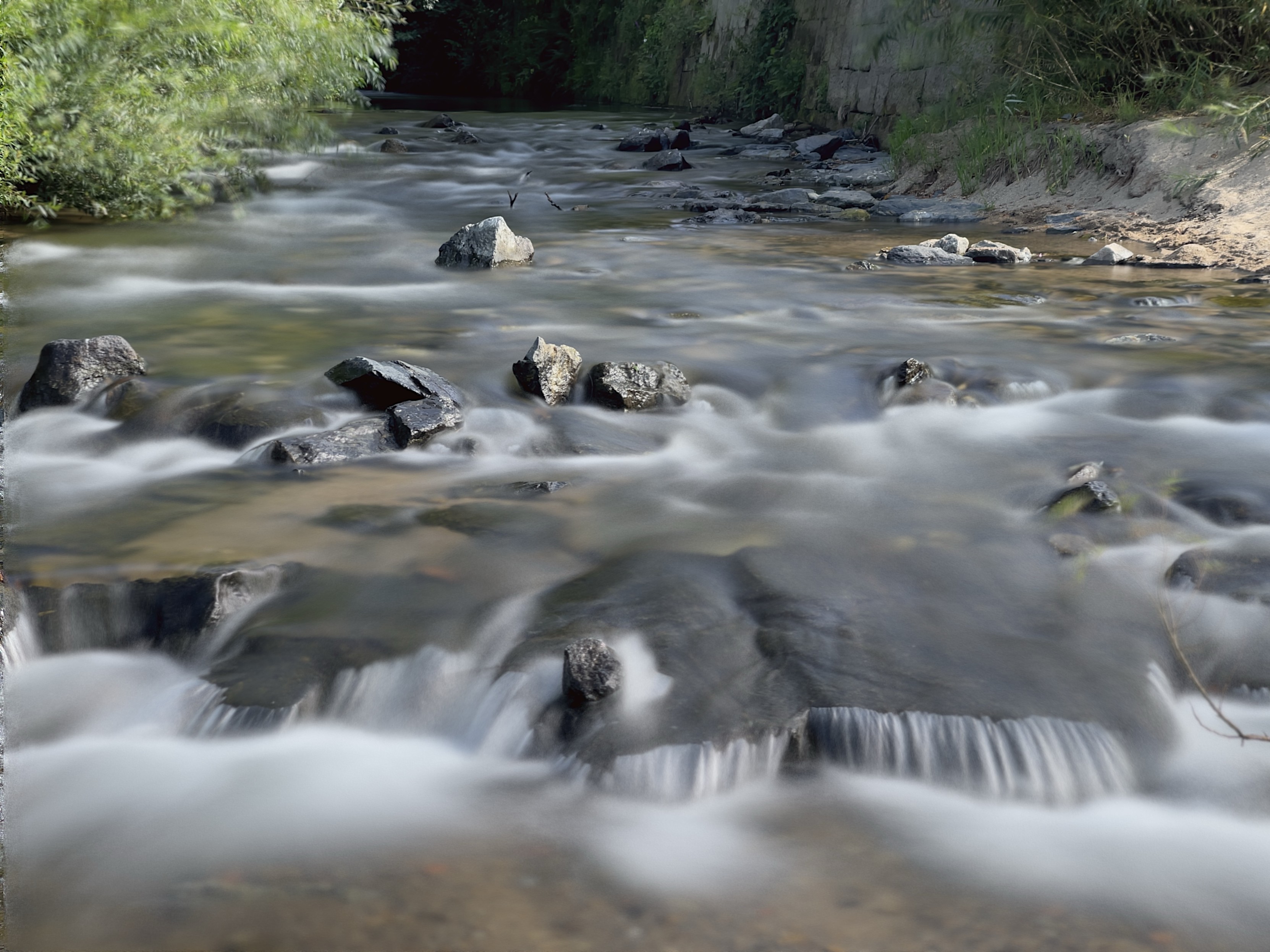 A serene stream with gently flowing water over rocks, surrounded by lush greenery.