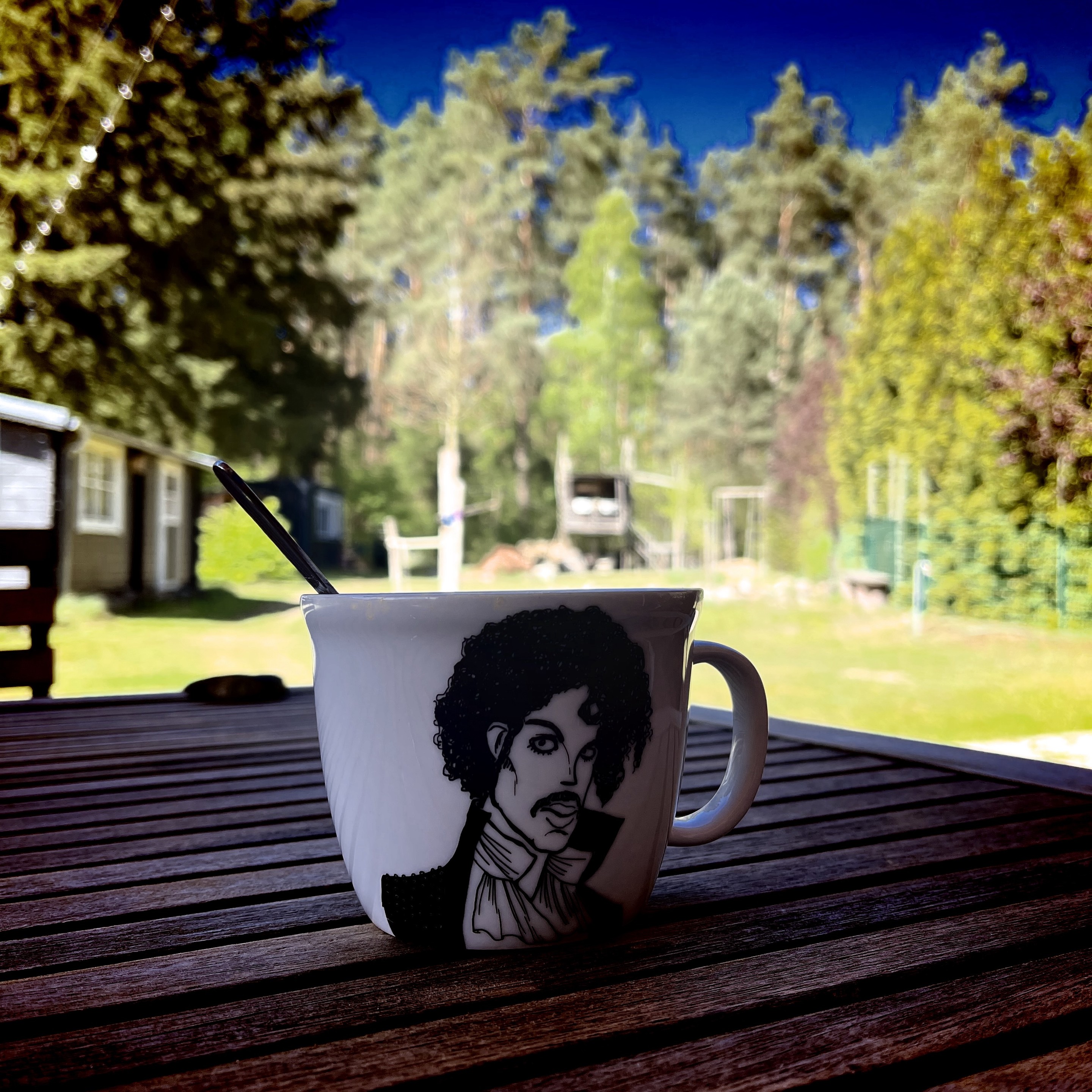 Ein Kaffeebecher mit einem stilisierten Porträt von Prince auf einem Holztisch, im Freien mit Bäumen und einer Hütte im Hintergrund.