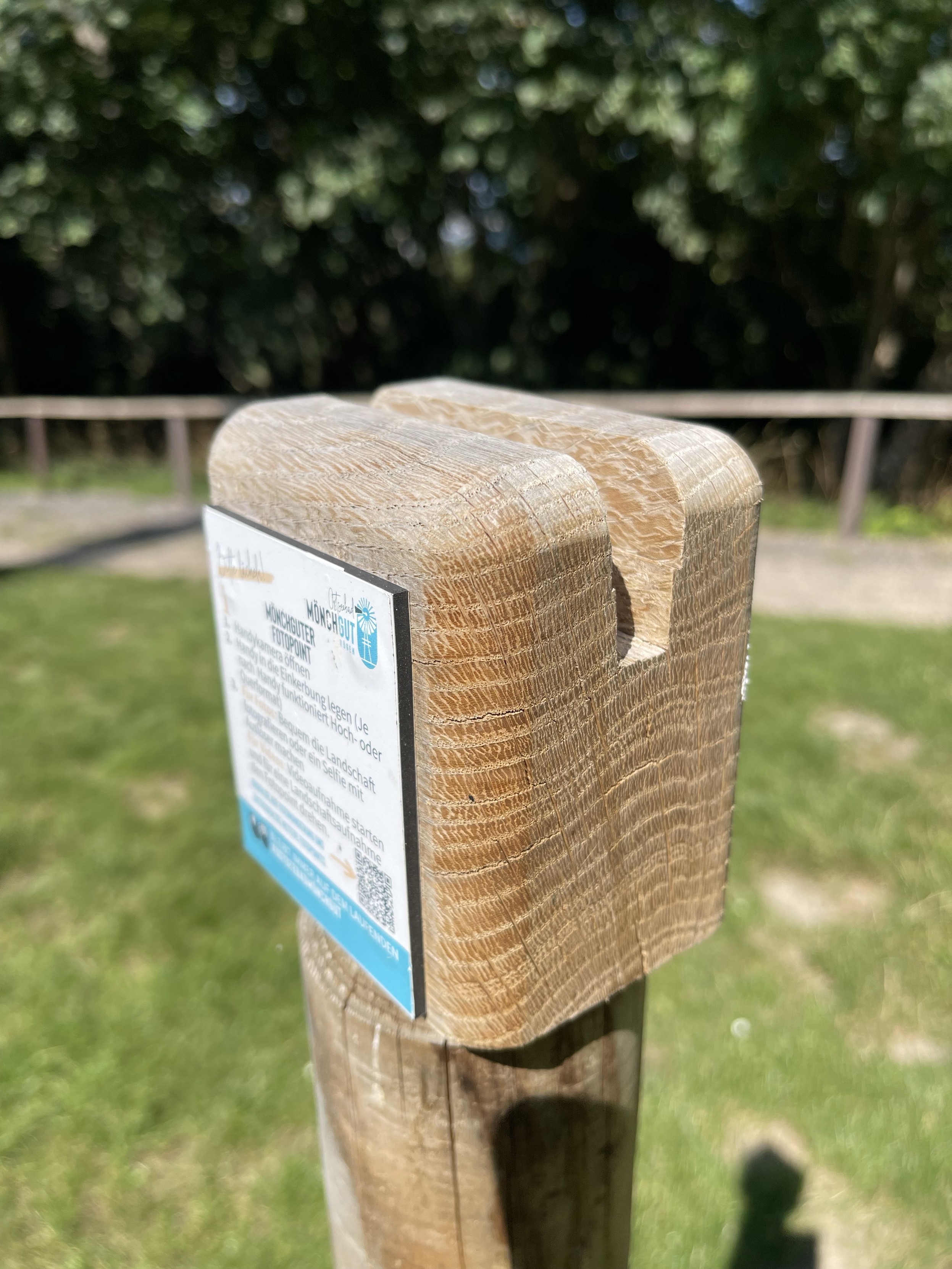 Holzpfosten mit einem Holzblock mit Schlitz für ein Handy mit einem kleinen Schild, das sich in einer grasbewachsenen Außenumgebung mit Bäumen im Hintergrund befindet.