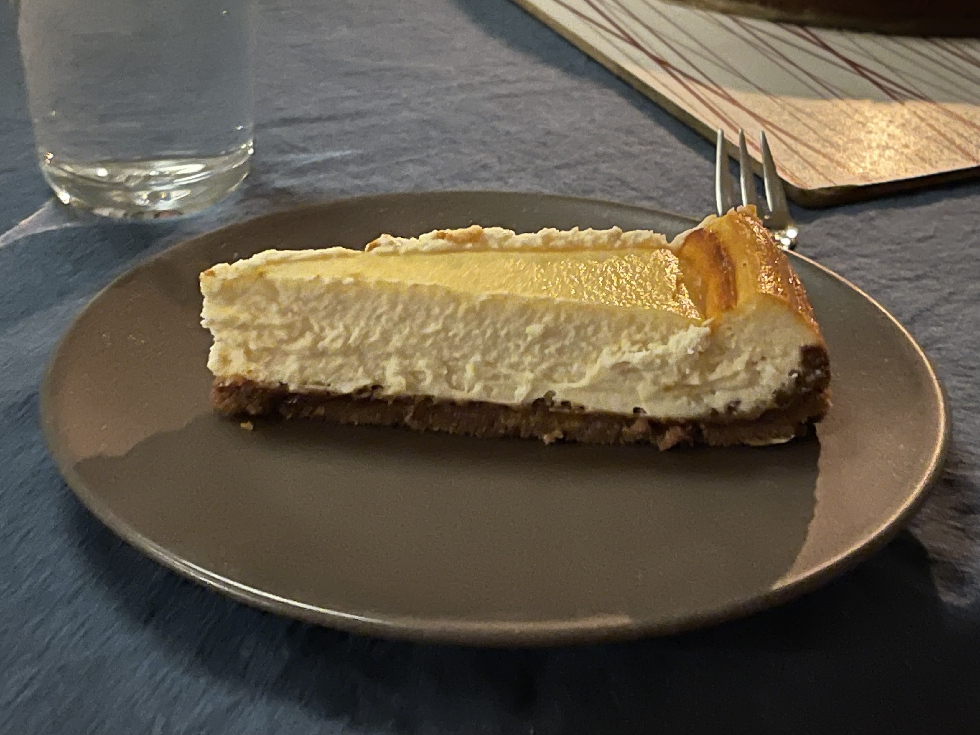 A slice of cheesecake on a brown plate, with a glass of water and a napkin with utensils in the background.