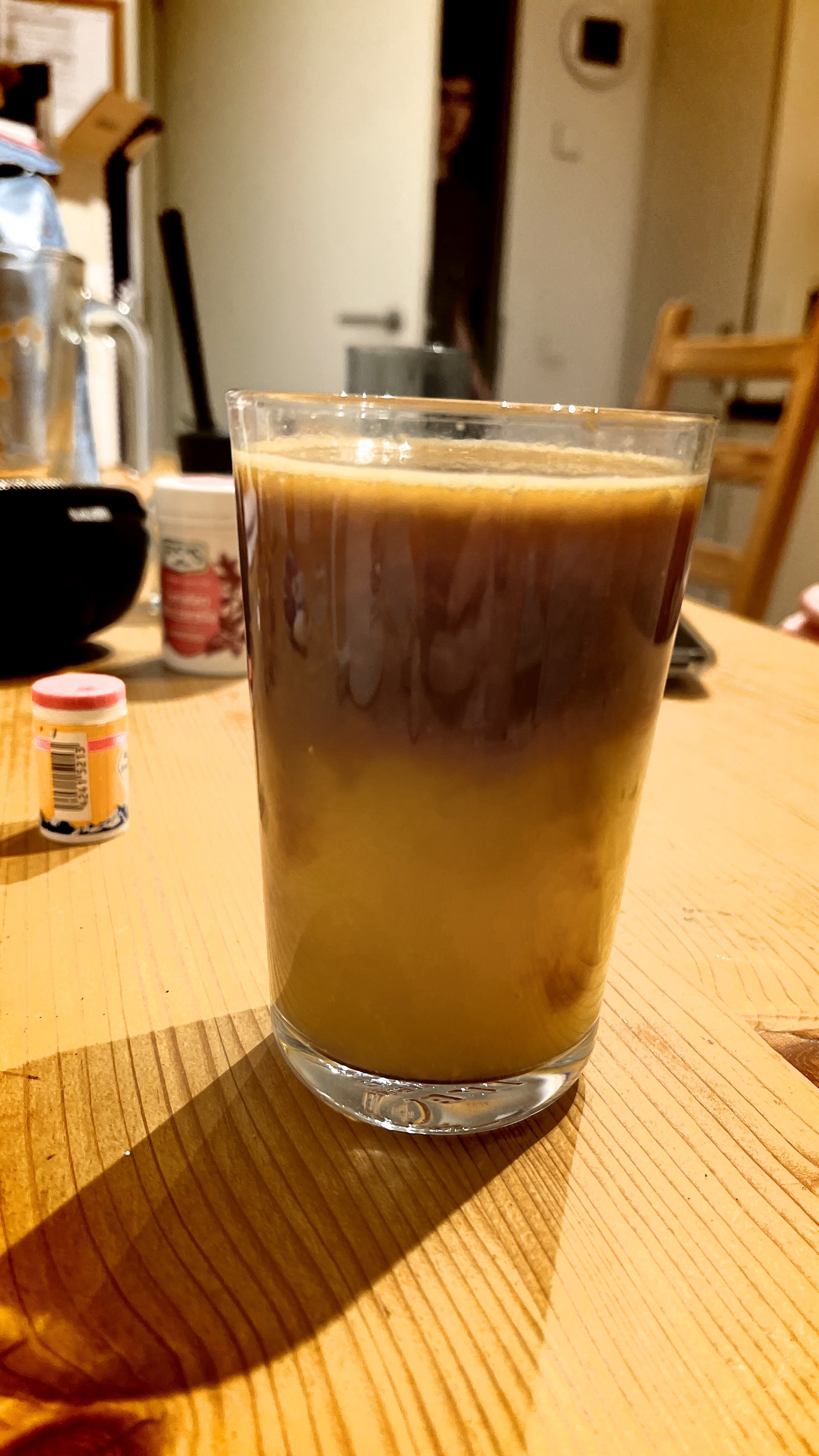 A glass of orange espresso. The beverage is layered. The orange juice is at the lower two thirds and the espresso floating atop. The background is blurry and showing a kitchen table. 