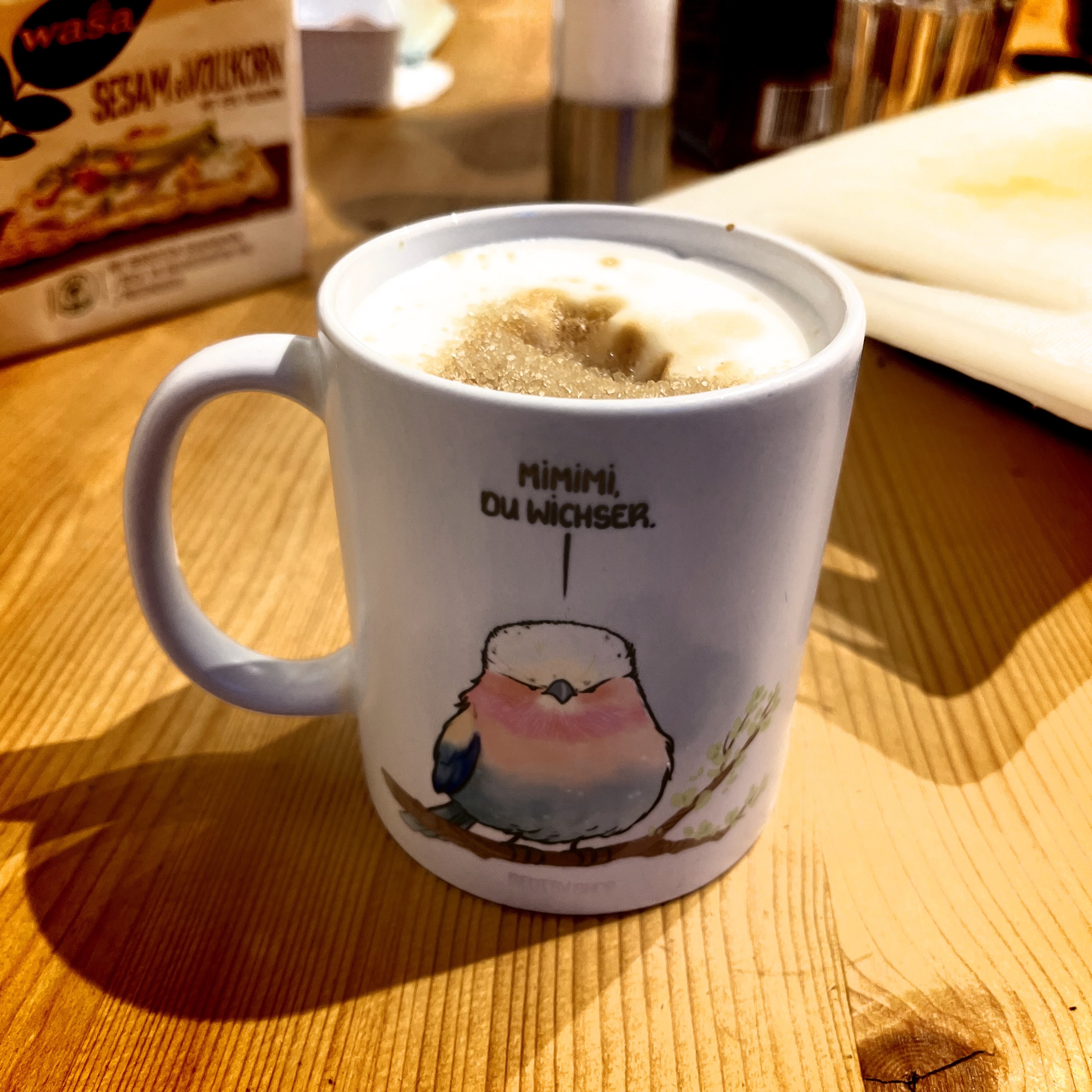 Eine Tasse Kaffee mit Schaum obenauf, mit einem Cartoon-Vogel und Text, auf einer hölzernen Oberfläche mit einer Schachtel Wasa-Knäckebrot im Hintergrund. Der Vogel sagt: Mimimi du Wichser