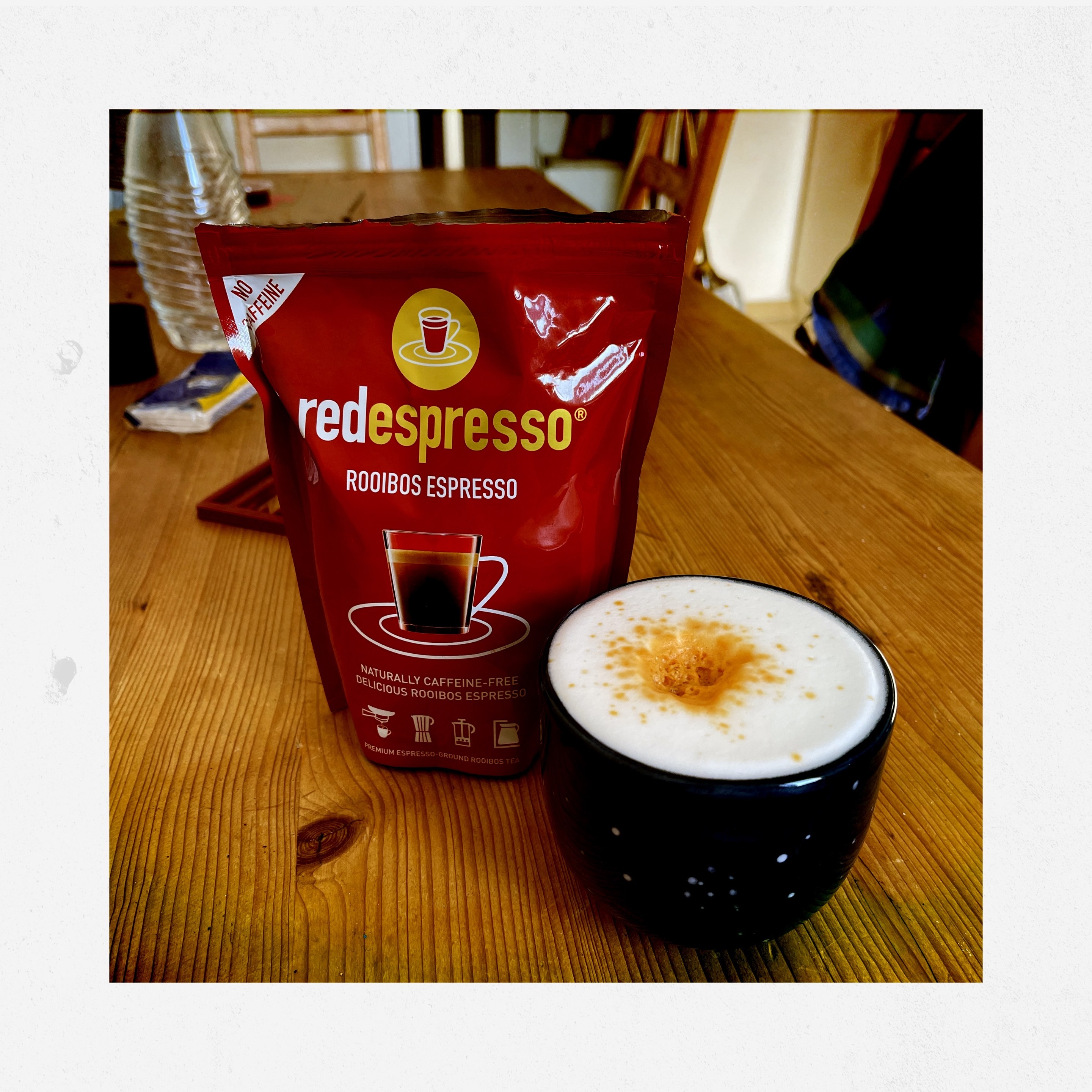 A red bag of redespresso (rooibos espresso) standing next to a dark blue cup with frothed milk and some orange/light brown sprinkles and n top. Both standing on a wooden table. 