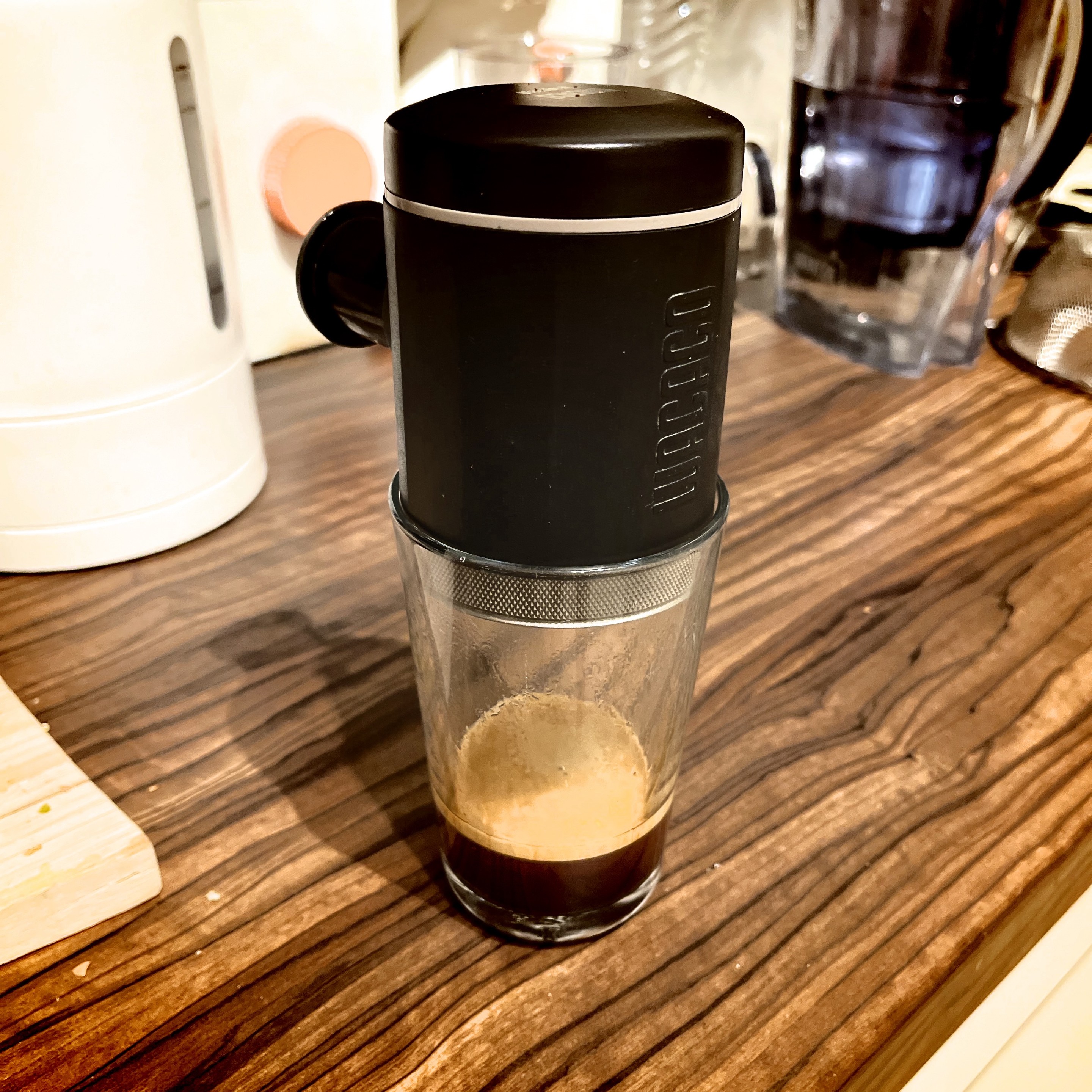 A portable coffee maker (Wacaco Nanopresso) sitting onto a glass on a wooden countertop with a small amount of coffee concentrate a.k.a. Espresso 