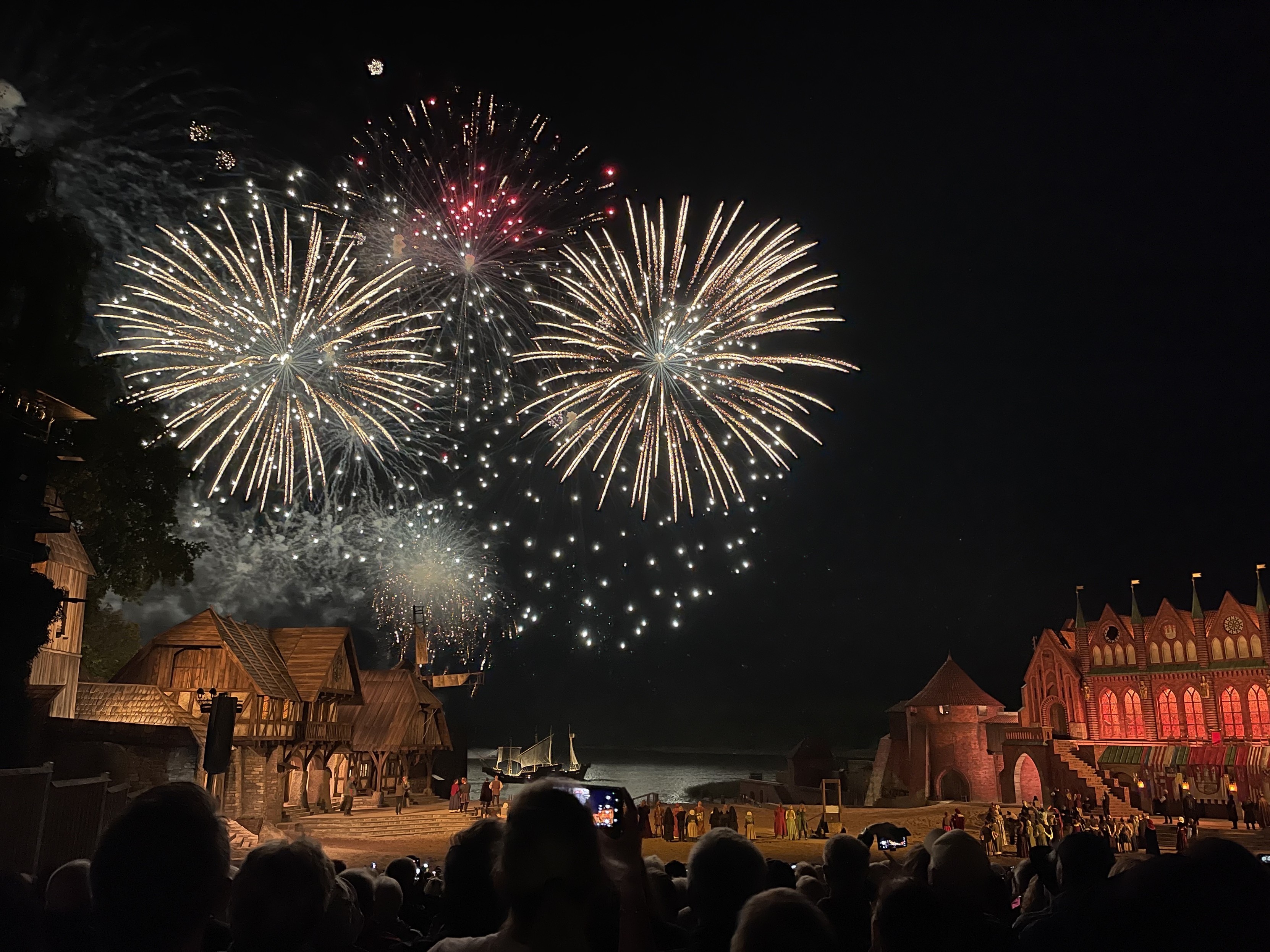 Feuerwerk, das den Nachthimmel über einer Menge beleuchtet, die eine Nachstellung oder Veranstaltung in der Nähe einer Uferpromenade mit Gebäuden im mittelalterlichen Stil und einem großen, kunstvoll beleuchteten Gebäude beobachtet.