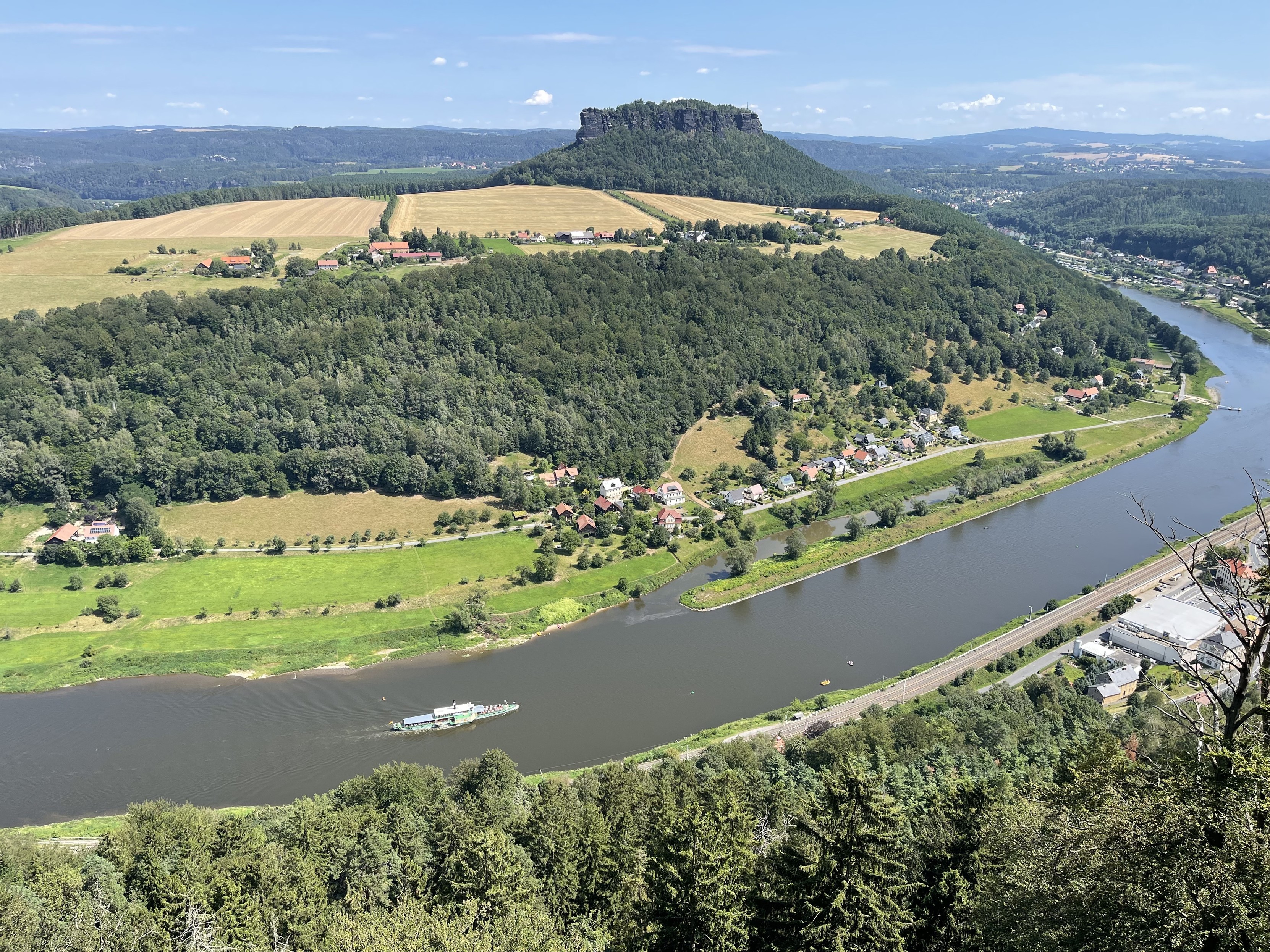 Scenic landscape of a river winding through a lush valley with hills, farmland, and scattered houses, viewed from a higher elevation.