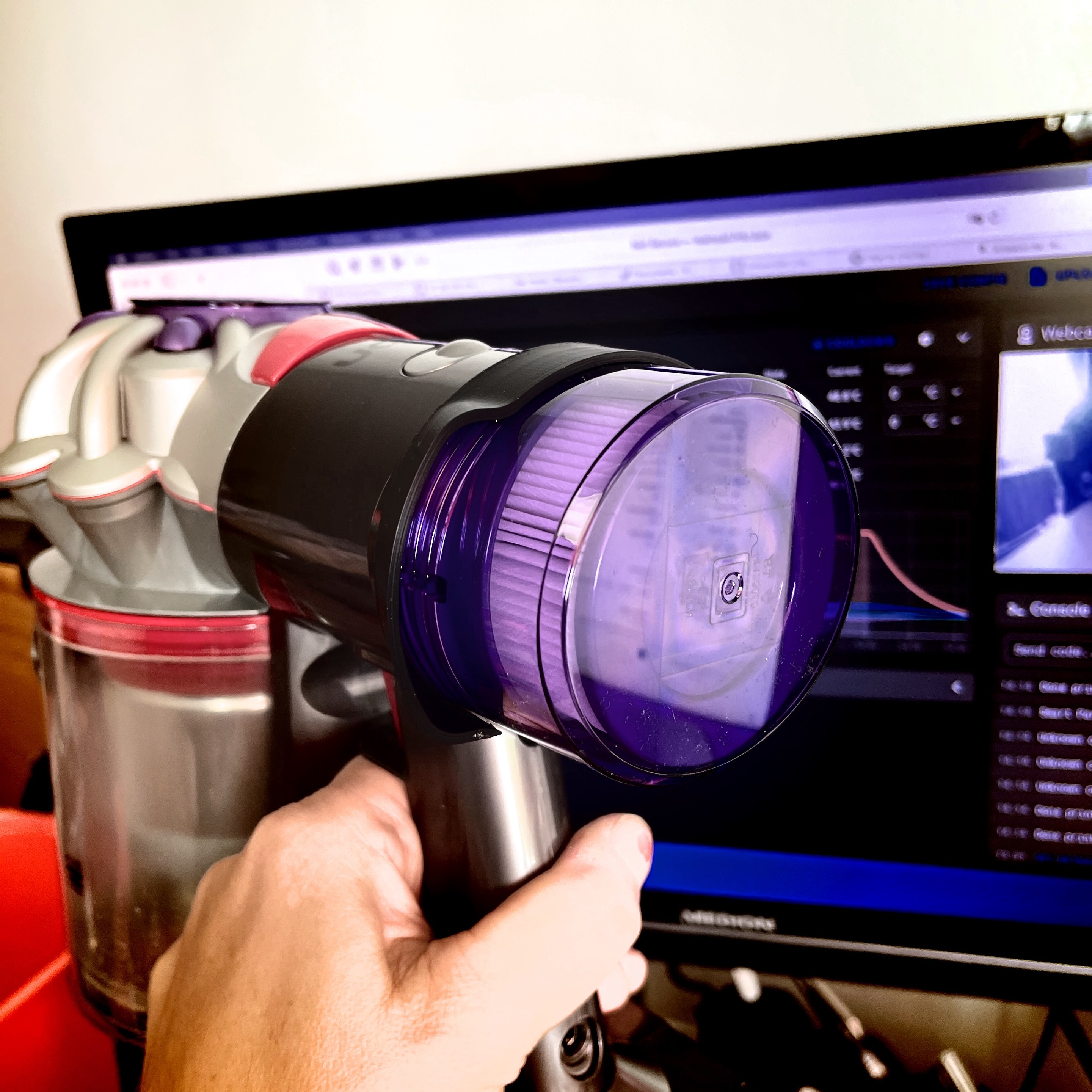 A hand holding a cordless vacuum cleaner with a computer screen in the background. The back of the purple filter housing now holds a 3D printed air diversion lid. 
