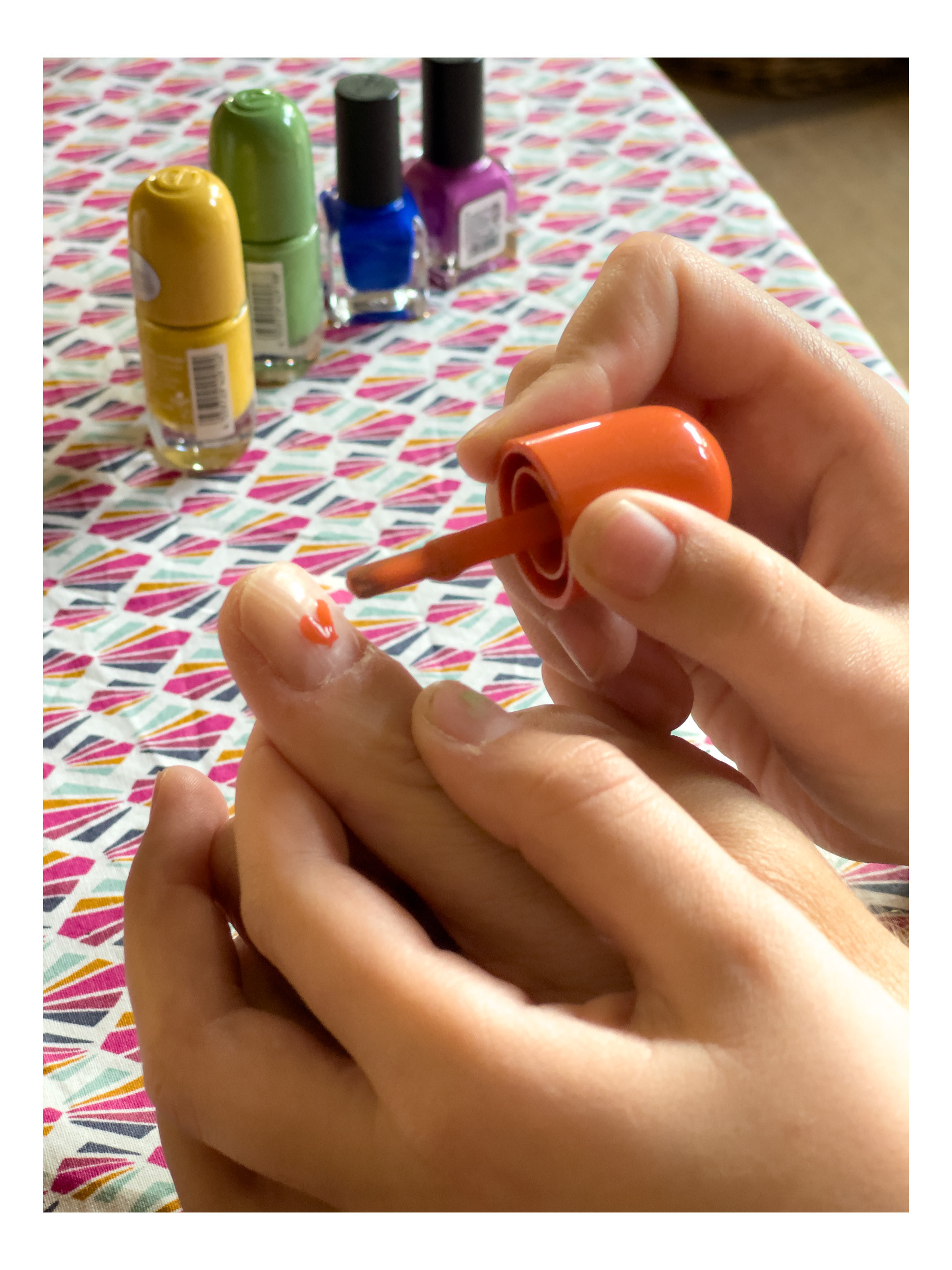 a right hand getting its nails painted by two hands of a child. Orange color is applied to the thumb at the moment. Colors yellow, green, blue , velvet waiting in the background on the table. 