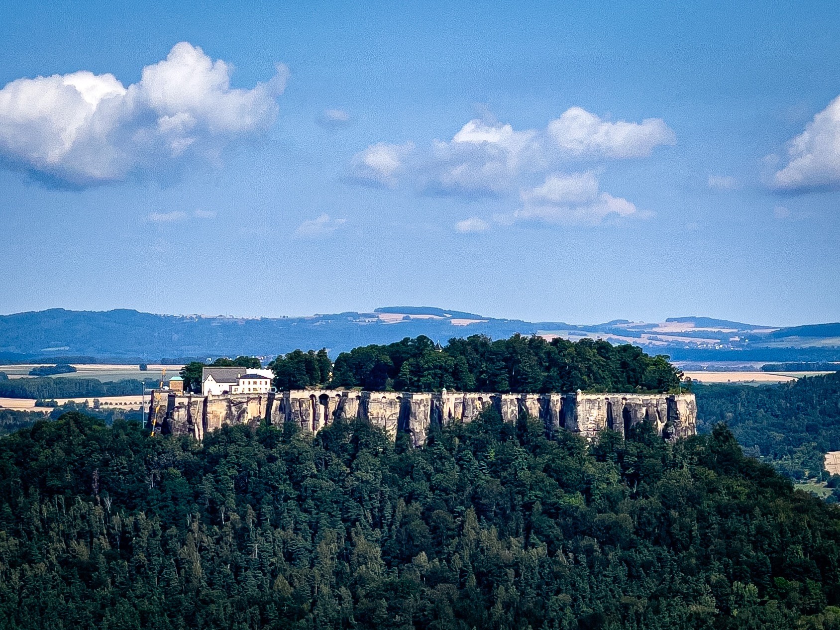  Festung, umgeben von dichtem Wald, unter einem blauen Himmel mit verstreuten Wolken. Entfernte Hügel und Felder im Hintergrund.

Das Schloss ist die Festung Königstein