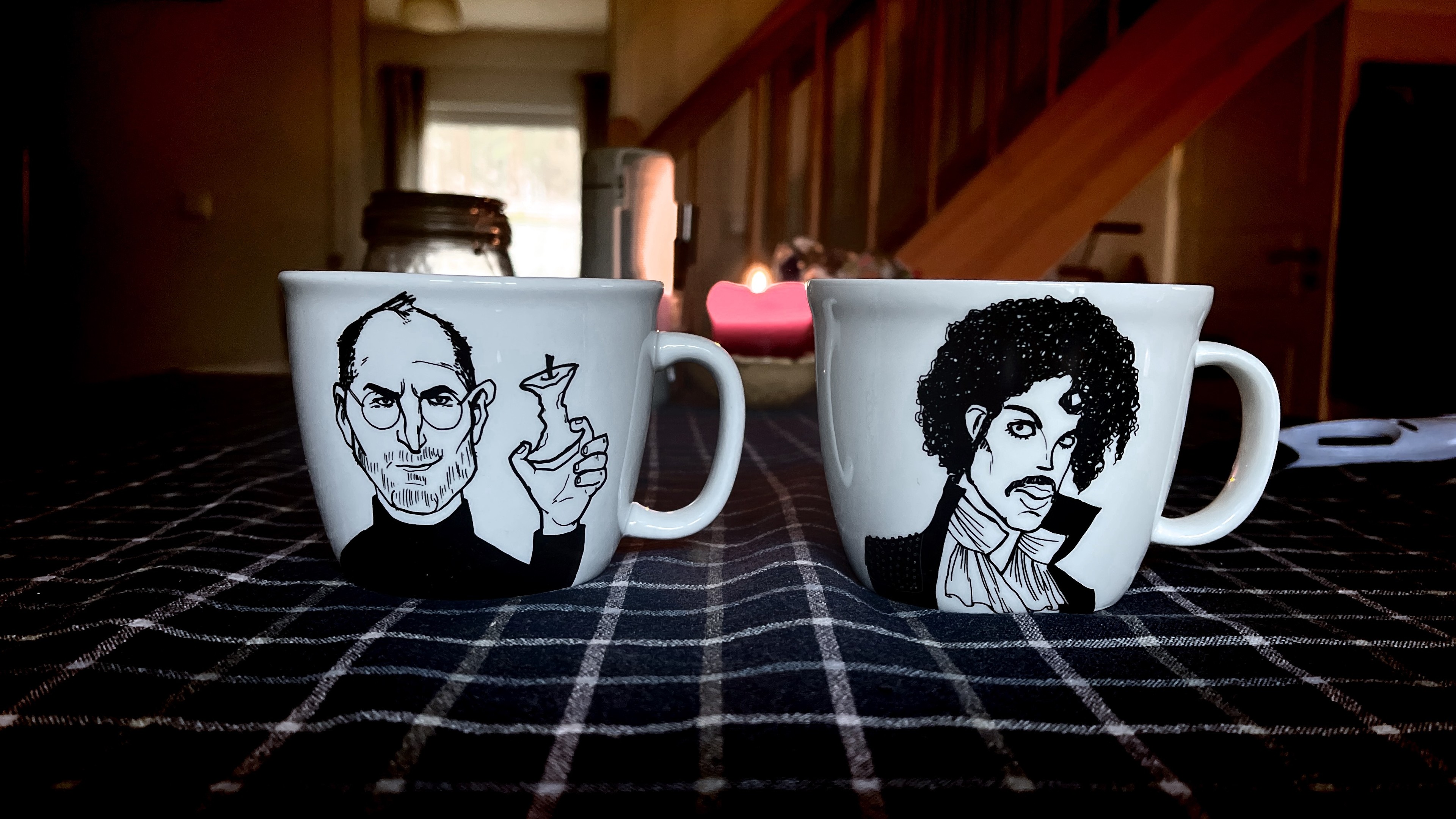 Two coffee mugs with illustrated portraits on a checkered surface, with a cozy room in the background.